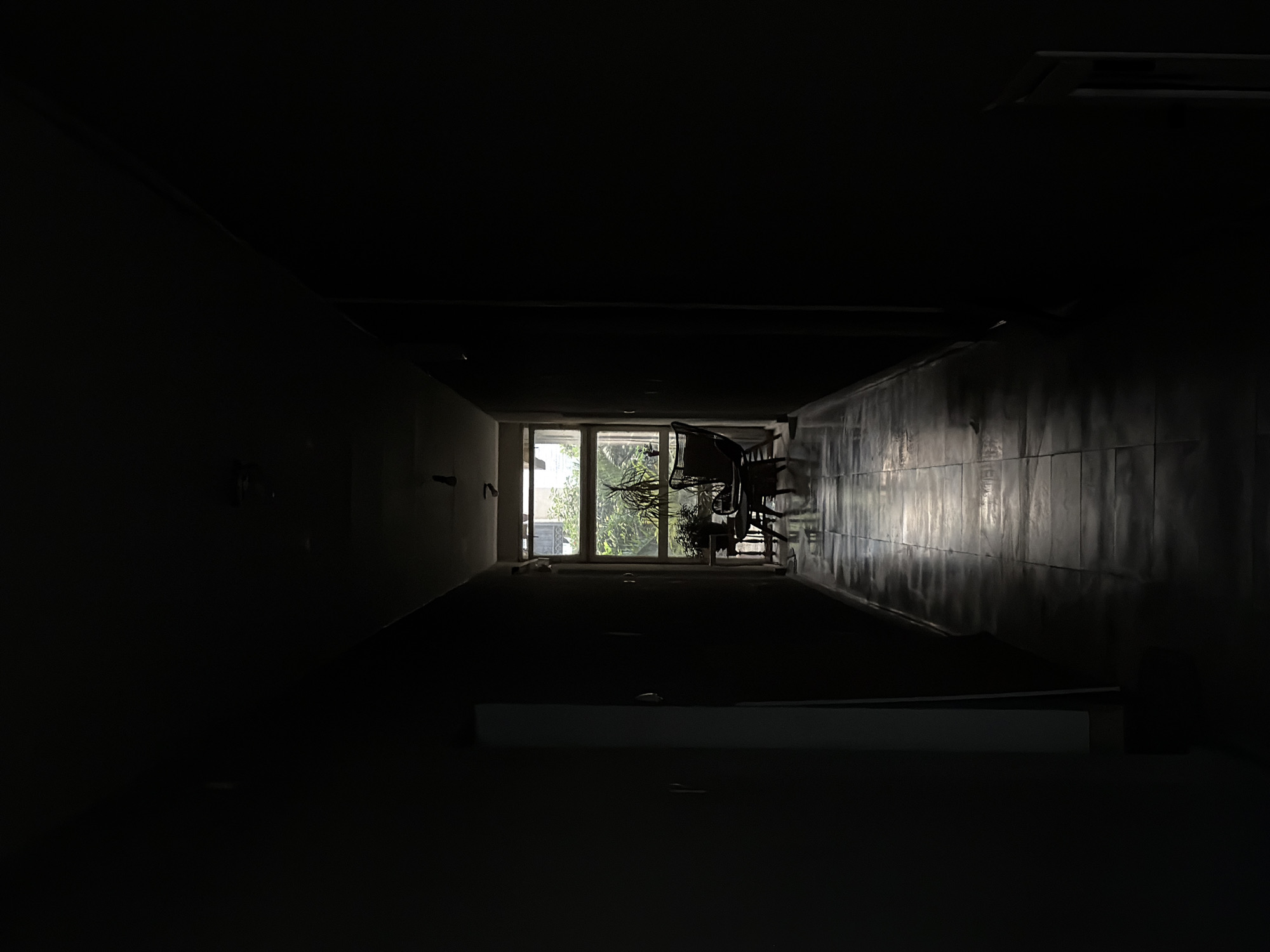 The image shows a long, dark, narrow hallway with a bright window at the far end. The hallway has tile walls, and the floor appears to be unlit. There are several dark shapes of chairs and what appears to be an exercise bike silhouetted against the light from the window at the end of the hallway.
The location in the city of Budva, Montenegro, is unknown.