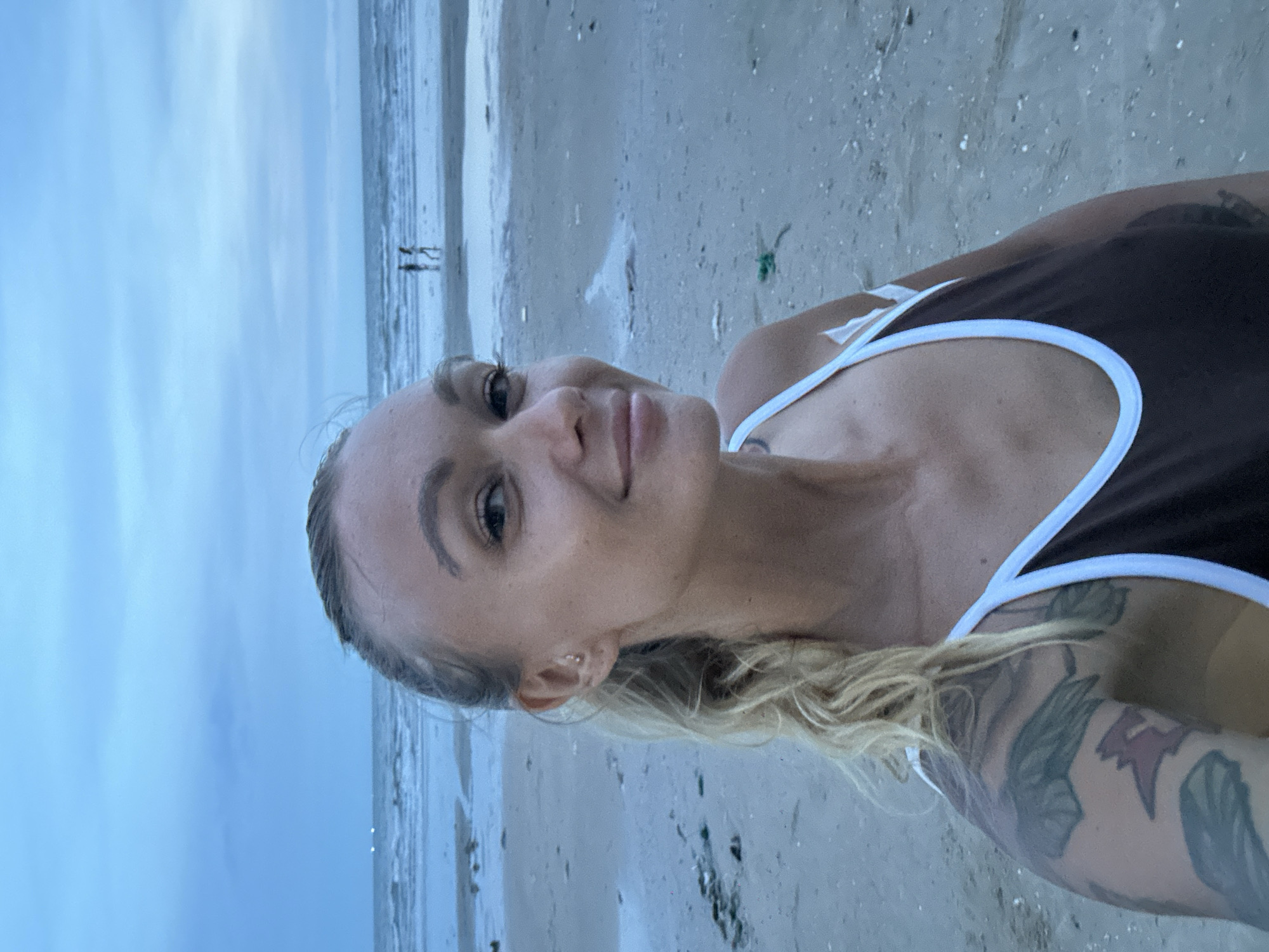 An outdoor, eye-level, medium shot captures a blonde woman with fair skin and a ponytail as she takes a selfie on a beach in Mũi Né, Vietnam. She is wearing a brown and white-trimmed swimsuit and has tattoos on her left arm. Her gaze is directed towards the camera, and she has a slight smile. The background shows a sandy beach with scattered debris and a tranquil ocean with a cloudy, blue sky overhead. Two figures can be seen walking in the distance along the shoreline.