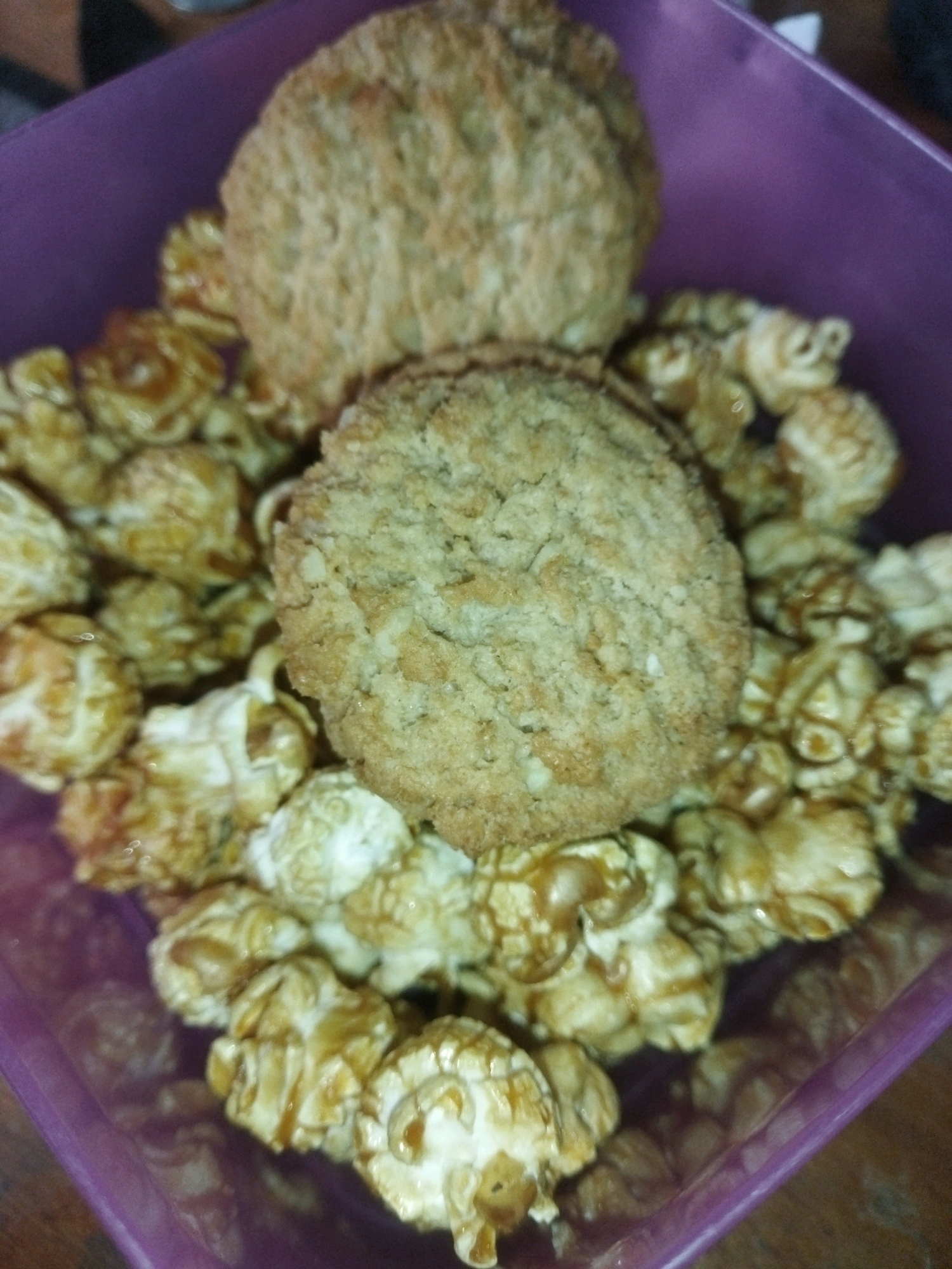 Two cookies are placed on top of caramel popcorn in a purple container. The cookies are round and appear to be oatmeal cookies. The caramel popcorn is golden brown and sticky, with some pieces clumped together. The container is translucent and purple, with a slightly rough texture. The image is taken from a high angle, looking down into the container. The background is blurred but appears to be a wooden surface. There are no people or any indication of a specific location in Tarai Bangun, Indonesia.