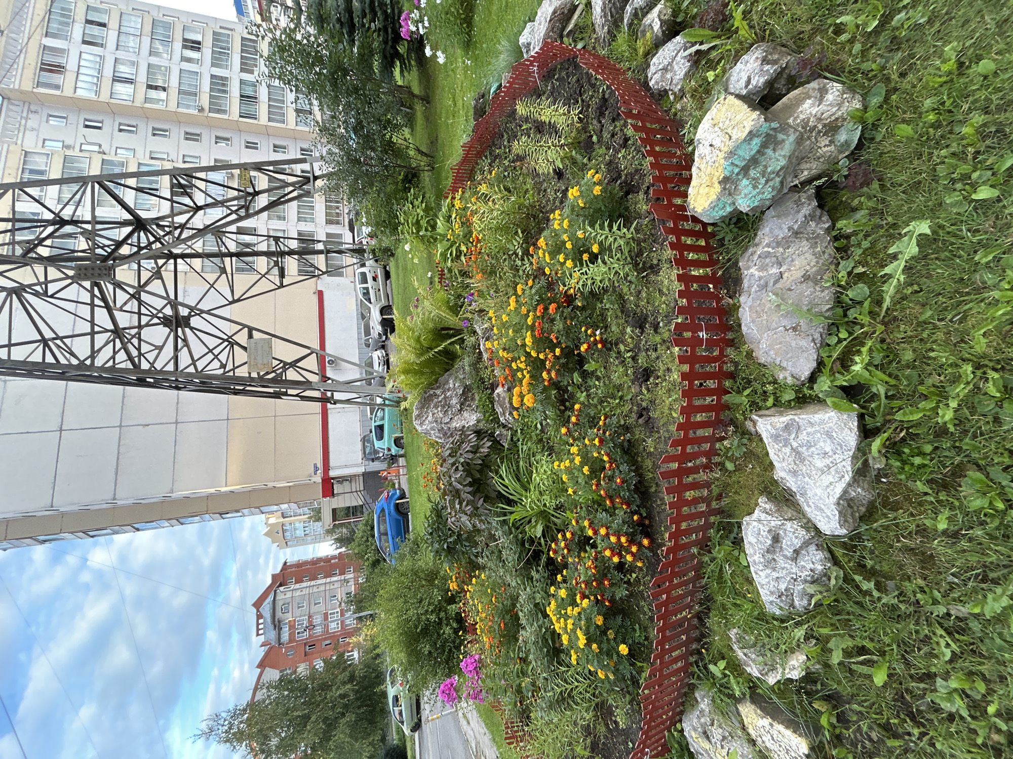 The image shows a flower bed in front of a multi-story apartment building in Tomsk, Russia. The flower bed is circular and surrounded by a red wooden fence. It is filled with a variety of colorful flowers, including yellow and orange marigolds, and some pink and purple flowers. Several large rocks are placed around the flower bed, some of which are painted with green and yellow accents. In the background, there is a large metal tower structure, and several cars are parked on the street. The sky is cloudy.