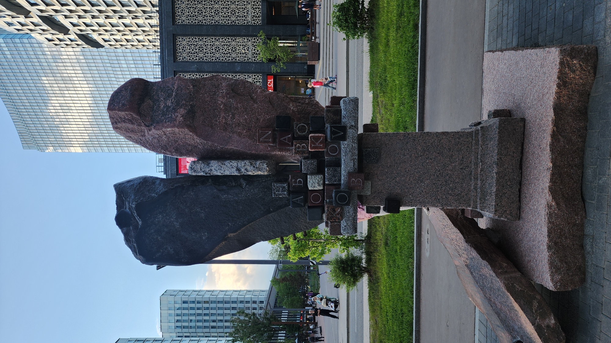 The image depicts a monument to Vladimir Nabokov in Moscow, Russia. The sculpture consists of two large, rough-hewn stones, one dark gray and the other reddish-brown, connected by a column of granite blocks with letters. The name "Nabokov" is spelled out in both Cyrillic and Latin alphabets. The monument is situated on a walkway near a green lawn and a paved area. In the background, modern buildings and pedestrians can be seen. The sky is visible in the upper left and lower right corners of the image.