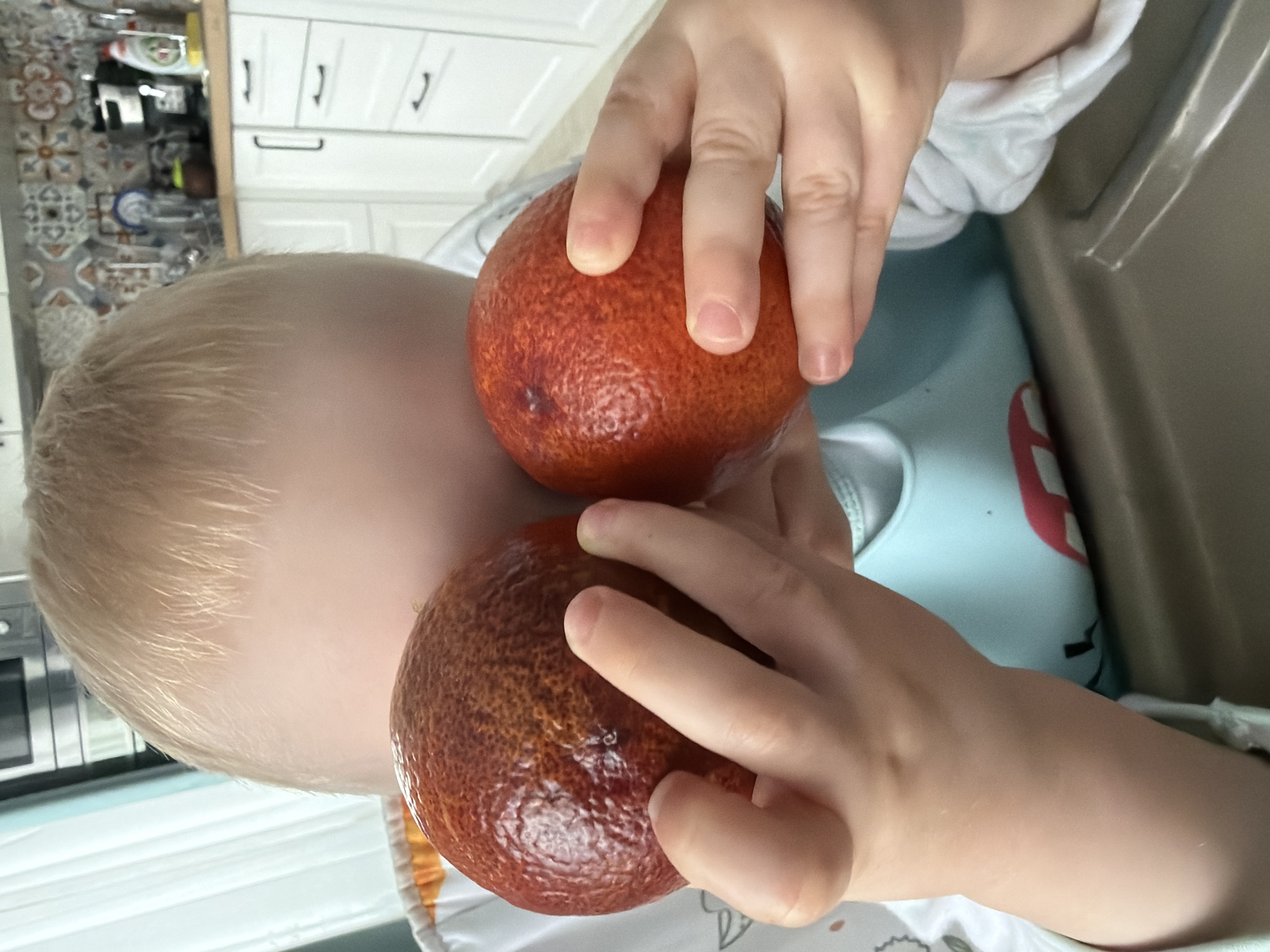 A child with blonde hair holds two blood oranges in their hands. The child is seated, possibly in a high chair, wearing a light blue top. The background includes white kitchen cabinets, tiled backsplash, and hints of kitchenware.