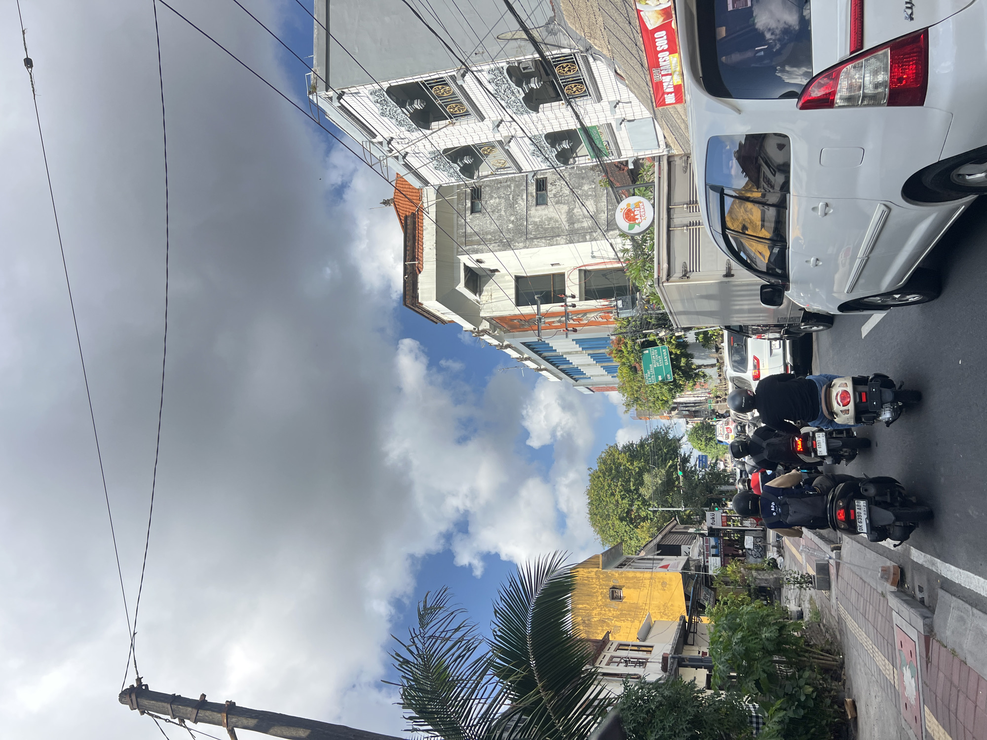 The image depicts a street scene in Denpasar, Indonesia, likely during daylight hours with scattered clouds in the sky. The perspective is from the middle of a traffic lane, looking forward. In the foreground, several vehicles are present, including a white SUV on the right and multiple motorcycles in front of it. The motorcycles are carrying riders, some with passengers, all wearing helmets. The traffic appears to be moving slowly or stopped.
The environment consists of a street with a paved sidewalk on the left, lined with buildings. On the left side of the street, there are palm trees and lush greenery. Buildings visible include a prominent yellow structure and several multi-story buildings with balconies and ornate architectural details. Some buildings appear to be commercial establishments, with signage visible, though text is mostly illegible. Overhead power lines and a wooden utility pole are present on the left side of the image. The road surface is asphalt, with painted lane markers. The overall impression is of a busy urban environment in Denpasar.