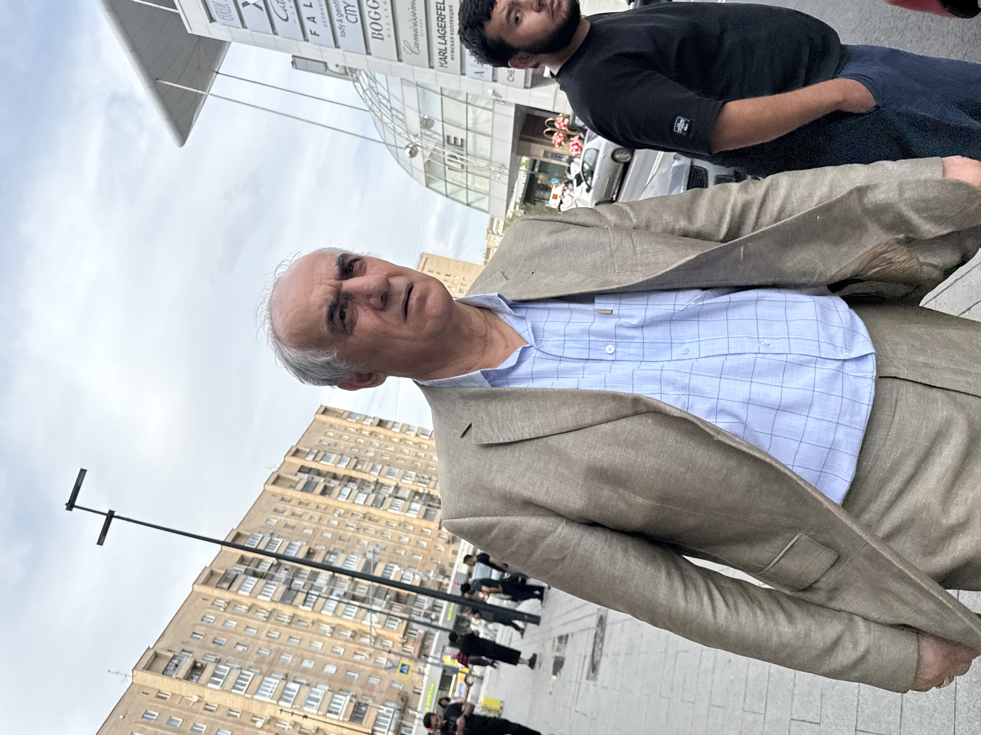 The image captures a street scene in Moscow, Russia, with a focus on two men in the foreground. The man on the left is older, with grey hair, and is wearing a light brown suit with a light blue checkered shirt. He is looking towards the right, with a neutral expression. The man on the right, younger with dark hair and a beard, is wearing a black t-shirt and dark pants, with his hands in his pockets. He is also looking towards the right. Behind them, a modern building with glass architecture and signs is visible, including one for "LIME" and other brands. To the left, a tall residential building with many windows stands against a cloudy sky. The street in the background shows other pedestrians and vehicles, indicating a public urban setting. The overall atmosphere is that of a typical day in a bustling city.