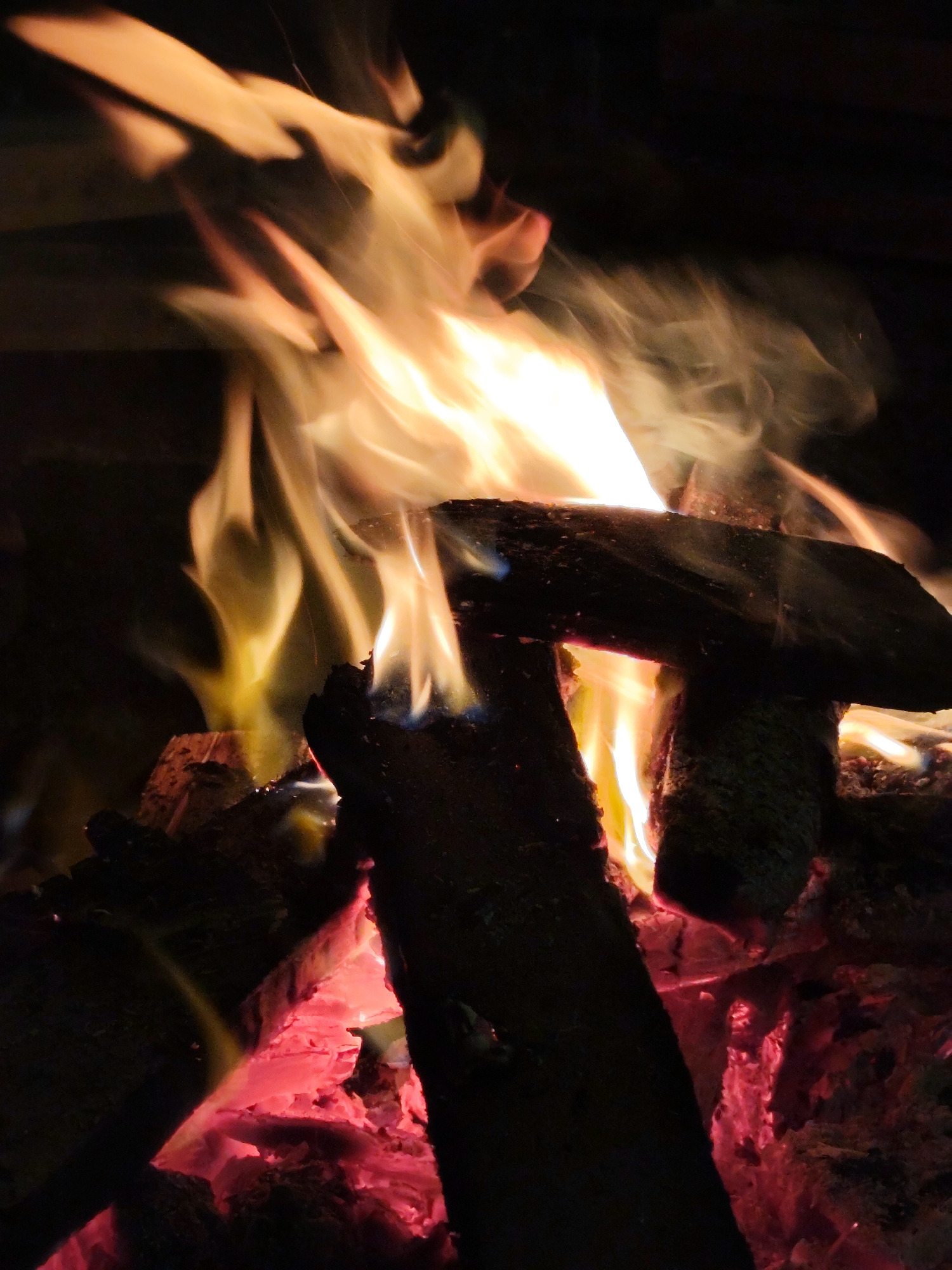 The image shows a close-up view of a bonfire in a dark setting. Several pieces of wood are stacked, some of which are burning, creating flames of varying colors from yellow to orange. The embers beneath the wood are glowing red. No people or specific identifiable objects outside of the fire and wood are visible. Xuan Truong - Da Lat, Vietnam is not identifiable in the image.