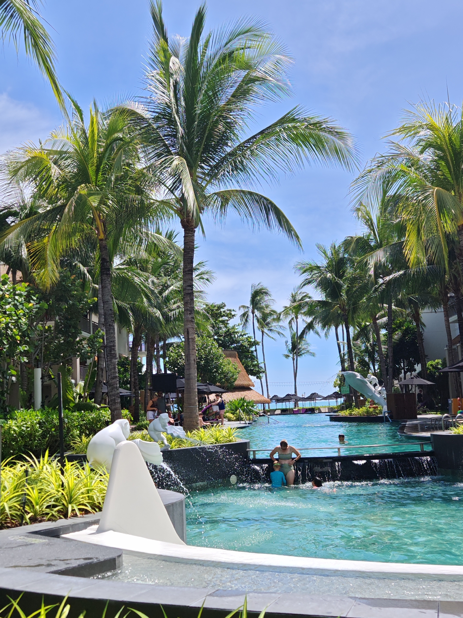 In the city of Baan Bo Phut, Thailand, a sunny day unfolds at a resort pool. Lush green palm trees line the background, their fronds reaching towards a clear blue sky. The pool itself is a vibrant turquoise, with a shallow area featuring a white abstract sculpture that appears to be a stylized wave or a bird. A small waterfall cascades into the pool from a dark-colored ledge.
In the foreground, two children and a woman are partially submerged in the water. The woman, wearing a green and white striped bikini, stands near the edge, facing away from the camera. One child, in a blue swimsuit, is in front of her, and another child is further in the pool. Another person is visible swimming in the deeper part of the pool.
In the mid-ground, to the right, a white dolphin-shaped slide is integrated into the pool's edge, with a person visible near its top. Umbrellas and lounge chairs are set up on the pool deck, and in the far distance, a sandy beach and calm ocean are visible under the bright sun. The scene is one of relaxation and family enjoyment in a tropical setting.