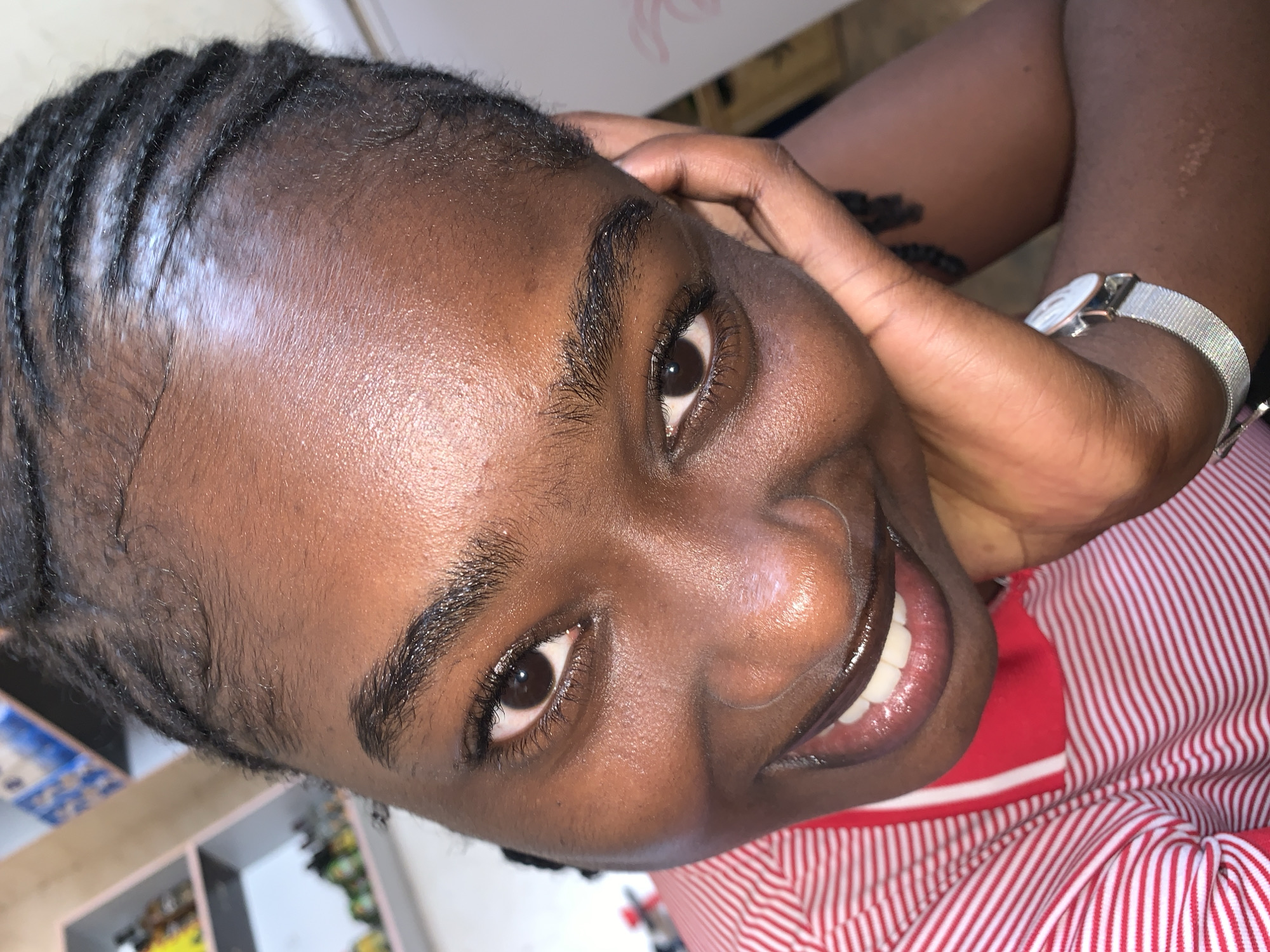 A close-up, angled portrait captures a young person, likely female, with dark skin, looking directly at the camera with a wide, friendly smile that reveals white teeth. Her dark hair is styled in cornrows, with some white residue visible along the partings on her scalp. Dark, well-defined eyebrows and eyelashes frame her brown eyes. A silver-toned watch with a mesh band and a light-colored face is worn on her left wrist, with her hand resting casually near her head. She is wearing a red and white striped top. The background is softly blurred, showing hints of shelves or storage units with various items, suggesting an indoor or semi-indoor setting. The person exudes happiness and engagement. The image is contextualized as being taken at New Stadium, Nigeria. No visible text is present in the image.