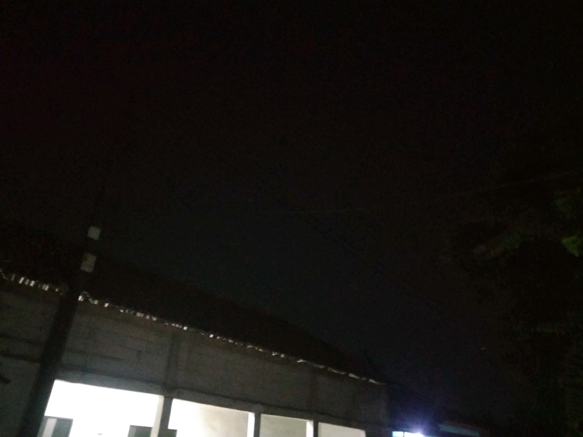 The image depicts a dark, nighttime scene, likely in the city of Suka Sari, Indonesia. The foreground shows the upper portion of a simple, weathered building with a corrugated metal roof. A dark, vertical pole, possibly a support structure, is visible on the left side of the image, partially obscuring the building's facade. The building appears to be under construction or unfinished, with exposed structural elements and open doorways or window frames, some of which are illuminated by artificial light, casting a bright white glow. The sky above is predominantly black, with a faint, possibly distant, light source. A few thin, horizontal lines, likely power lines or wires, are faintly visible against the dark sky. To the right, the dark foliage of trees can be seen, suggesting a natural element bordering the built environment. The overall impression is one of an outdoor, semi-developed area at night, with the building being the primary man-made structure.