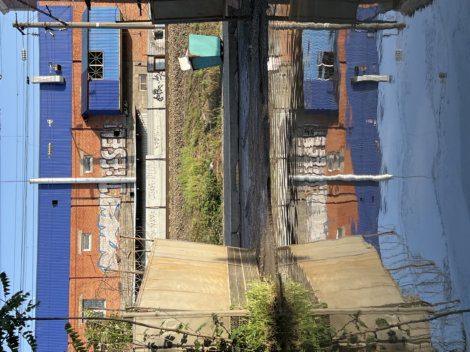 The image depicts an urban landscape with a reflection in water. On the left, there's a building with a blue corrugated metal roof and brick walls, partially covered with graffiti. A utility pole runs along the left edge. Adjacent to the building is a railway track with gravel and sparse vegetation. A small green structure is positioned beside the track. The reflection in the water mirrors the buildings and sky, with distortions due to ripples. The scene is possibly located in an industrial or urban fringe area.