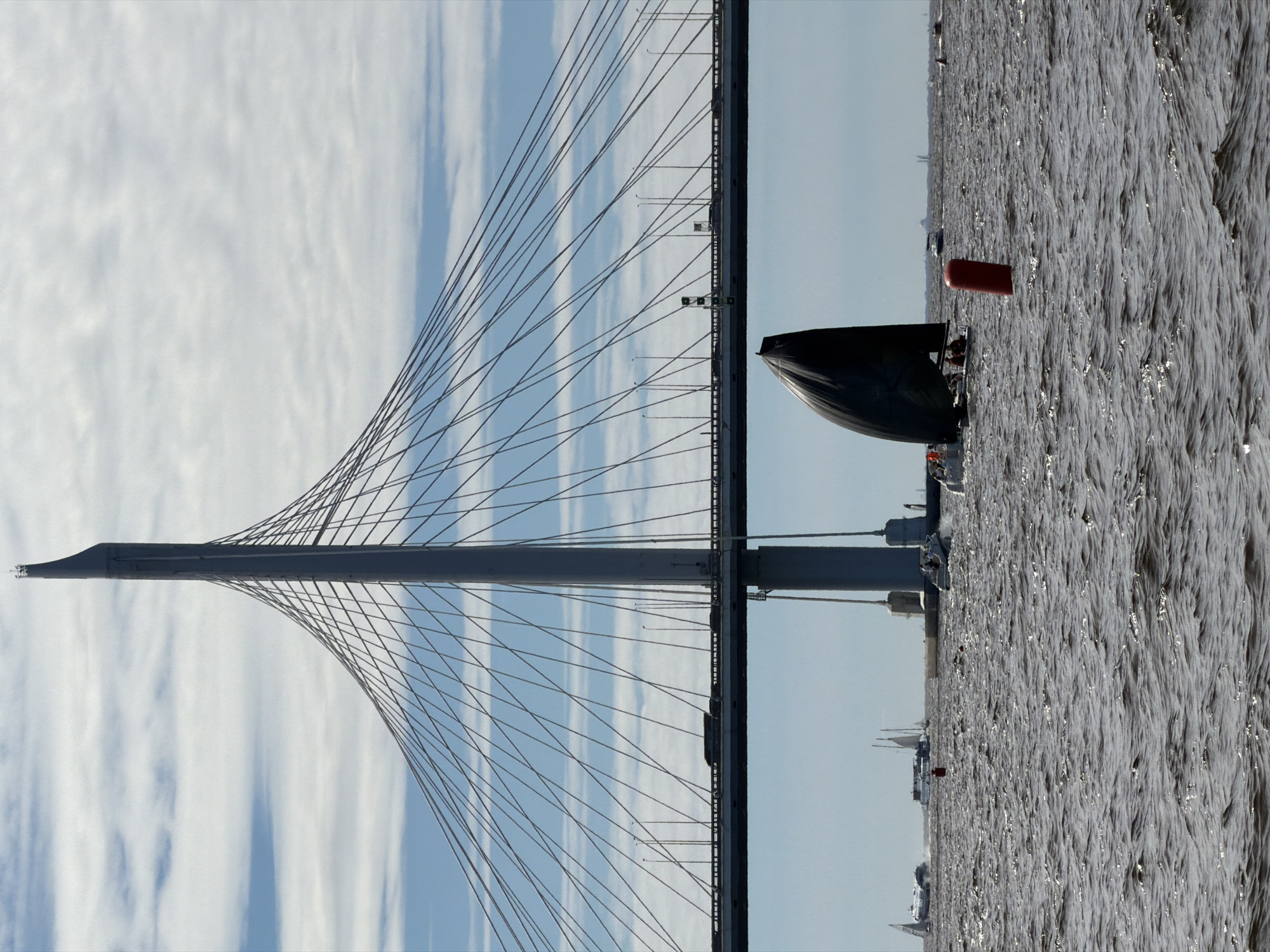 The image shows a wide shot of a body of water, likely a river or bay, under a partly cloudy sky. Dominating the background is a large, modern cable-stayed bridge, a prominent landmark of Saint Petersburg, Russia, likely the Western High-Speed Diameter. The bridge's towering pylon and intricate network of cables are clearly visible against the sky. A road surface with several vehicles can be seen crossing the bridge.
In the foreground and middle ground, several boats are on the water. The most prominent is a sailboat with a large, dark sail, tilted significantly, suggesting it is actively sailing or possibly capsizing. Several people are visible on the deck of this sailboat. Other smaller boats, including speedboats and a larger vessel resembling a yacht with multiple decks and masts, are scattered across the water, some appearing to be in motion. A red cylindrical buoy is also visible in the water near the sailboat. The water itself is choppy, with visible waves and sunlight reflecting off the surface. The overall scene suggests a day with active watercraft on a considerable body of water, under a major city bridge.
