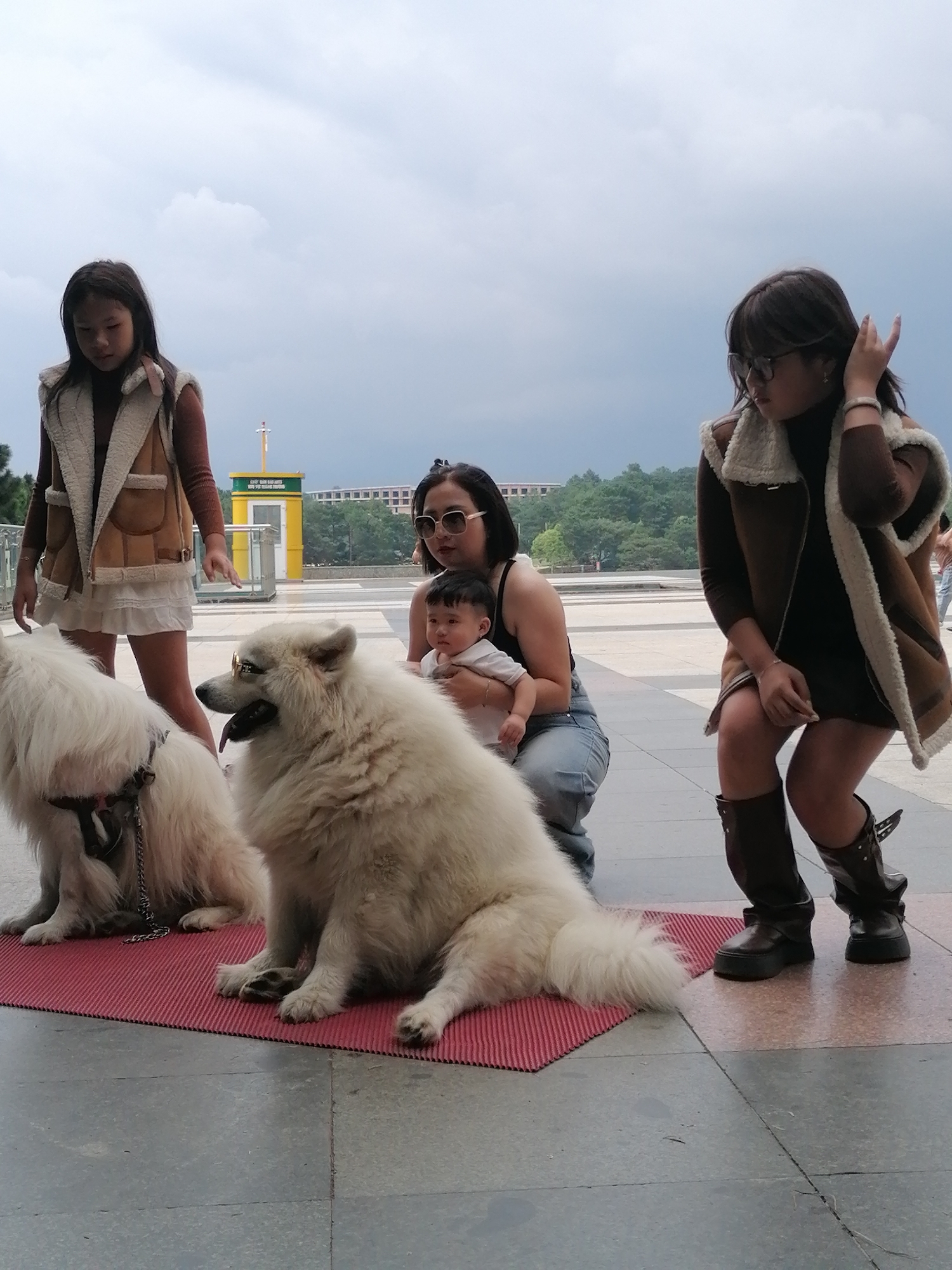 The image depicts an outdoor scene in the city of Da Lat, Vietnam, possibly in a public square or park, with overcast skies. In the foreground, two large, fluffy white Samoyed dogs are seated on a red mat. The dog on the right is wearing sunglasses. A woman is kneeling behind the dog on the right, holding a baby. To the left of the dogs, a young girl in a brown vest over a rust-colored long-sleeved shirt and white skirt stands looking down. To the right of the kneeling woman, another young girl or woman, wearing a similar brown vest over a dark outfit and dark boots, is partially visible. In the background, there is a yellow structure resembling an elevator or information booth, trees, and buildings. The overall atmosphere suggests a casual gathering.