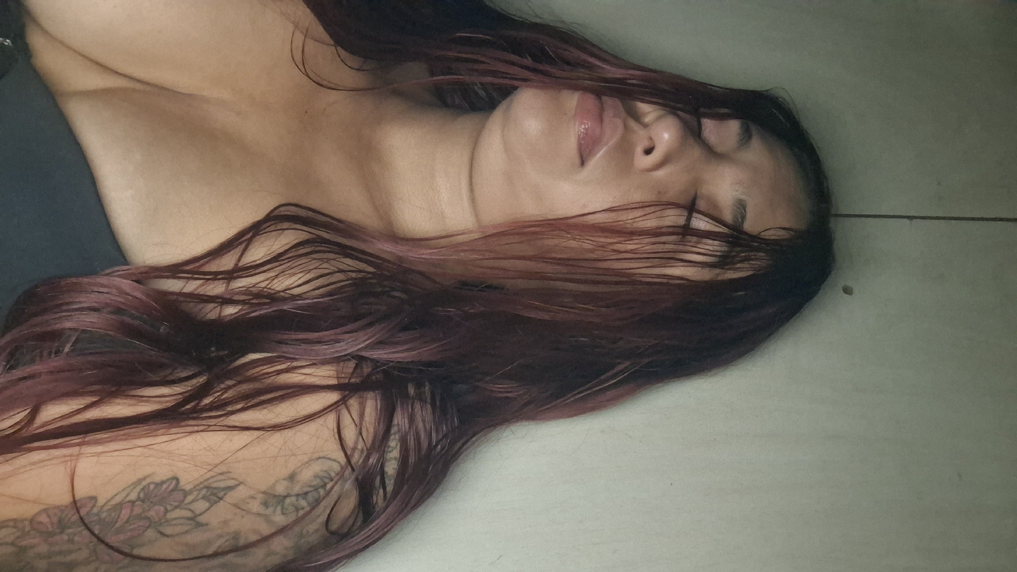 The image shows a close-up of a woman with her eyes closed, her head resting against a wall. Her hair is long and dark red, with some strands falling across her face. She is wearing a dark-colored garment that reveals her chest. There is a tattoo on her left shoulder. The background appears to be a plain, light-colored wall. The lighting is dim. The image does not provide any information about the city of Vallecito, Venezuela.