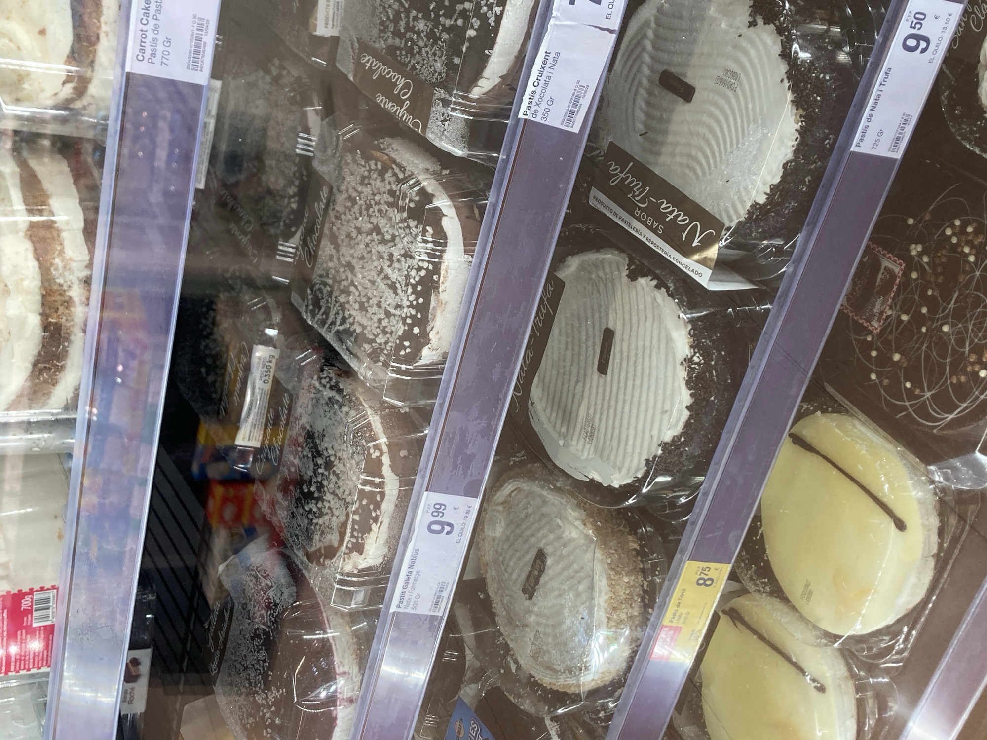 This media file is a close-up, slightly angled shot of a refrigerated display case in a supermarket, likely in Barcelona, Spain. The primary subjects are various packaged cakes and pastries. The products are presented in clear plastic packaging, allowing the viewer to see the desserts inside. The display is well-lit, typical of a retail environment.
Several distinct products are visible. On the left, a "Carrot Cake" (Pastís de Past) weighing 770g is prominently displayed, with layers of cake and cream. To its right, a "Pastís Cruixent de Xocolata i Nata" (Crunchy Chocolate and Cream Pastry) weighing 350g is visible, featuring a dark chocolate base and white cream topping, sprinkled with coconut. Further to the right, a "Pastís de Nata i Trufa" (Cream and Truffle Pastry) weighing 72.5g is shown, a lighter colored dessert with a dark chocolate drizzle. Another product, "Pastís Galeta Nabius Nata i Formatge" (Nabius Biscuit Pastry, Cream and Cheese) weighing 500g, is also visible. Towards the right side, there are yellow-colored pastries with a dark chocolate stripe, labeled as "Pastís de Torró" (Nougat Pastry). The price tags and product labels are visible in Spanish and Catalan, with prices like "9.99 €" and "9.50 €". The overall scene suggests a typical grocery store setting where customers can select pre-packaged desserts. There are no people visible in the frame.