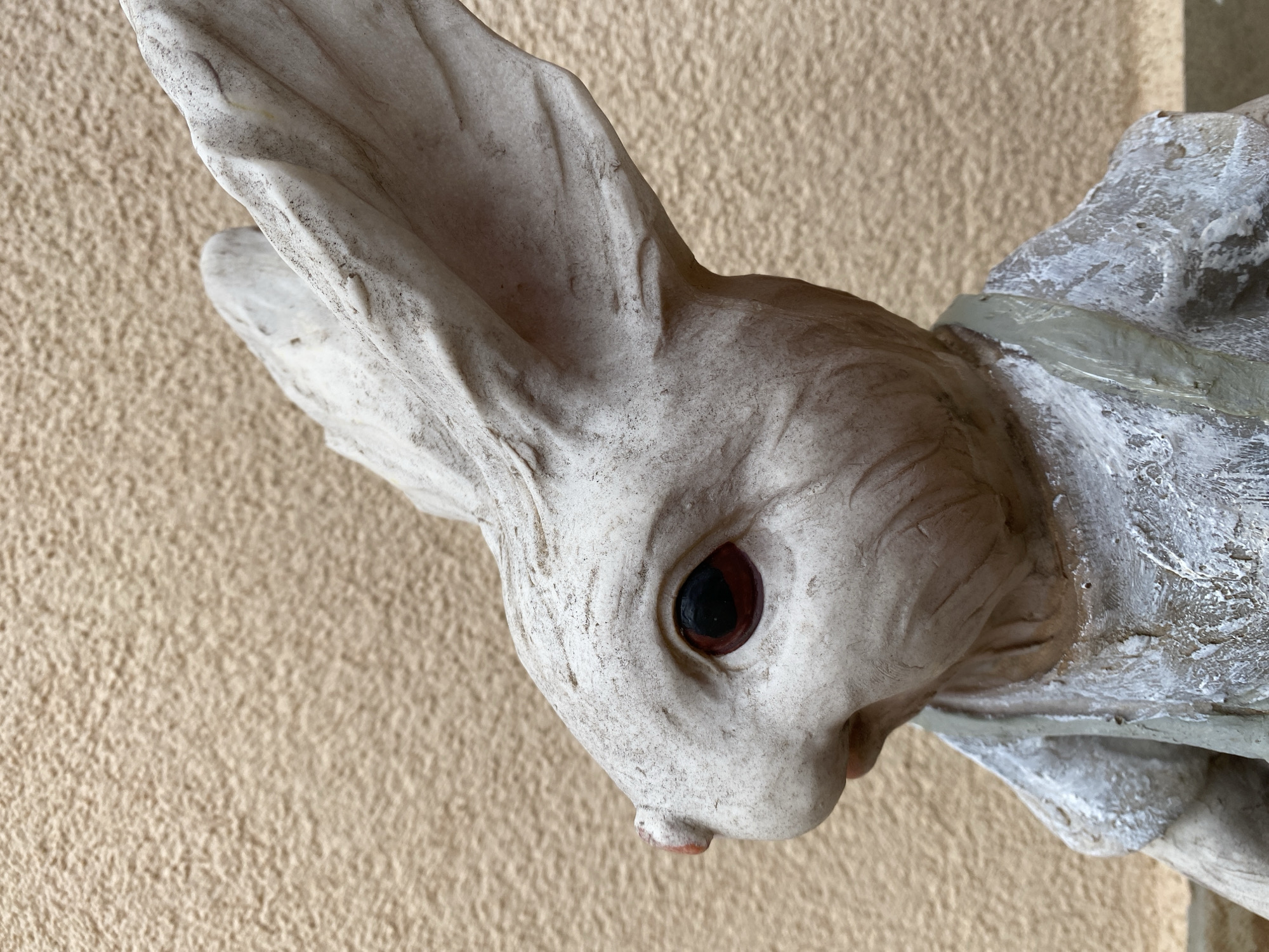 A close-up shot shows a weathered, light-colored statue of a rabbit's head and shoulder. The rabbit has one visible, dark eye. The statue's texture shows signs of age and wear. The background is a plain, light-tan wall. The location is unknown and not discernible from the image.