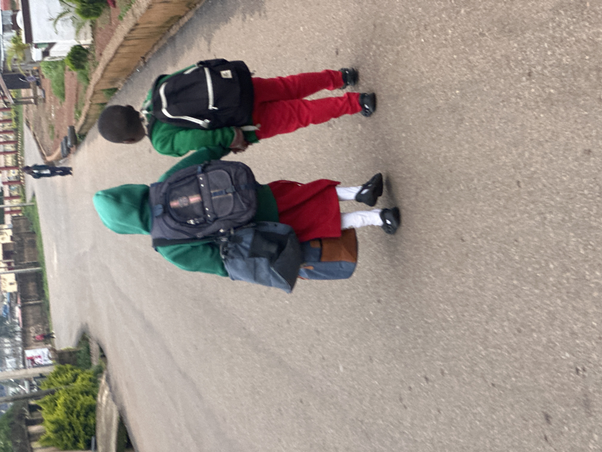 Two children, viewed from behind, walk hand-in-hand on a paved road. The child on the left wears a green hooded sweatshirt, a blue backpack, and a red skirt over white socks and black shoes. The child on the right wears a green sweatshirt, red pants, and black shoes. Both children have backpacks. The road appears to be in a town or city, with buildings and vegetation visible in the background. The scene is likely in Gura Top, Nigeria.