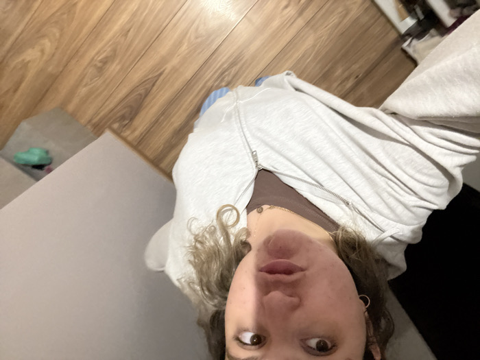 A person with light skin, wearing a gray zip-up hoodie over a brown top and gold necklaces, takes a selfie from a low angle. The background includes light brown wood flooring, a gray surface with a green object, and a dark area on the right. The person has brown eyes, curly blonde hair, and is wearing hoop earrings.