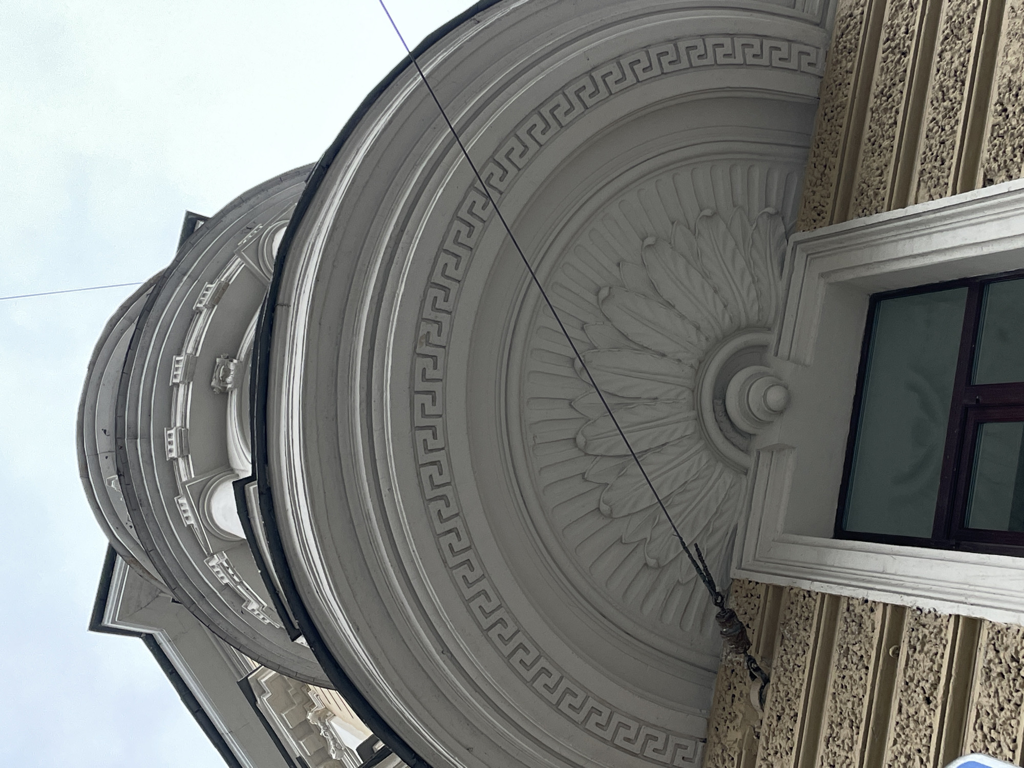 The image shows a low-angle view of an architectural detail of a building in Saint Petersburg, Russia. The building features a curved, decorative facade with neoclassical elements. There is a large, circular molding with a Greek key pattern surrounding a central rosette-like design. A window is visible to the right, with a dark frame. The wall beneath the window is textured with vertical lines. A cable or wire cuts across the frame from the top left to the lower right.