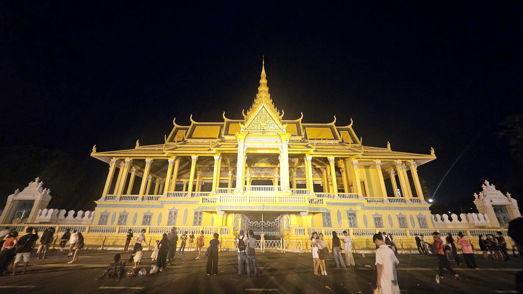 The image depicts the Royal Palace in Khan Daun Penh, Cambodia, at night. The ornate yellow structure, illuminated by artificial lights, dominates the center of the frame. Its distinctive tiered roofs and intricate carvings are visible against the dark night sky. A tall, pointed spire rises from the central building.
In front of the palace, a paved area, likely a street or plaza, is populated by a diverse group of people. They are engaged in various activities: some are standing and observing the palace, others are walking, and a few appear to be posing for photographs. The individuals are dressed in casual attire, with a mix of genders and ages visible. Some people are holding bags or belongings.
To the left and right of the main palace building, parts of a white, decorative wall with gates are also visible. The overall atmosphere suggests a public gathering, with people enjoying the nighttime view of the illuminated palace. The lighting creates a dramatic contrast between the bright palace and the dark surroundings.