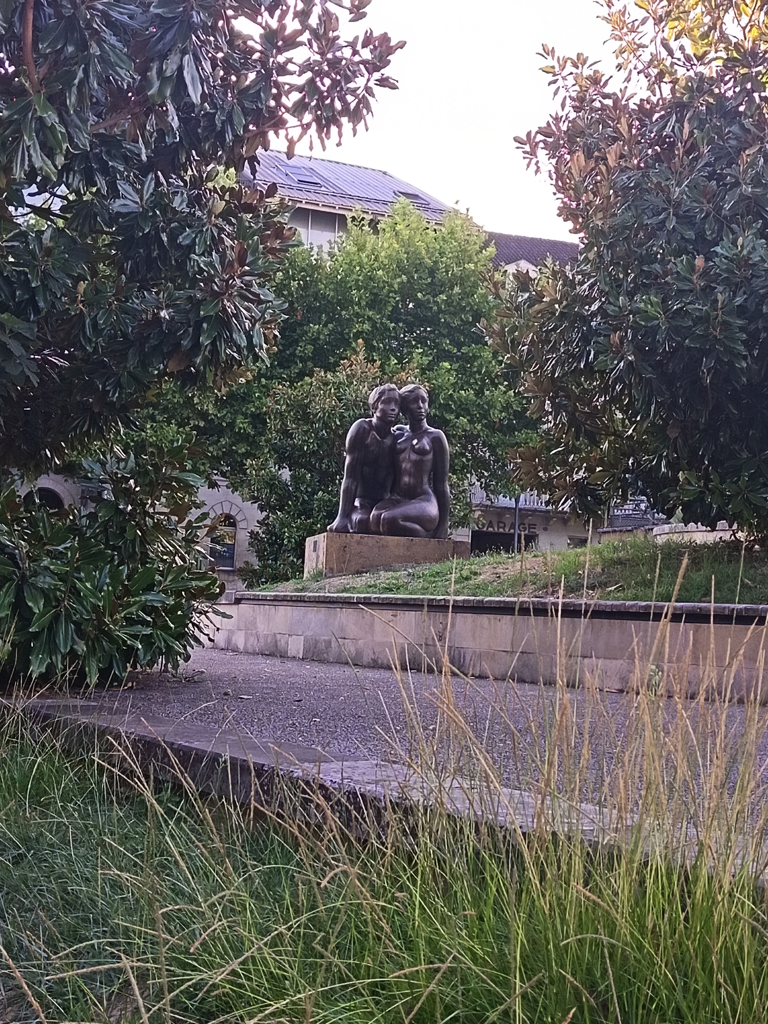 This image depicts a bronze sculpture of two figures embracing, situated in a park-like setting with lush green foliage. The sculpture is placed on a stone pedestal in the foreground, with tall grasses in front and mature trees framing the scene. In the background, a building with arched windows and a sloped roof is visible, along with a sign that reads "GARAGE". The overall impression is one of public art in an urban environment. The sculpture is located in Périgueux, France.