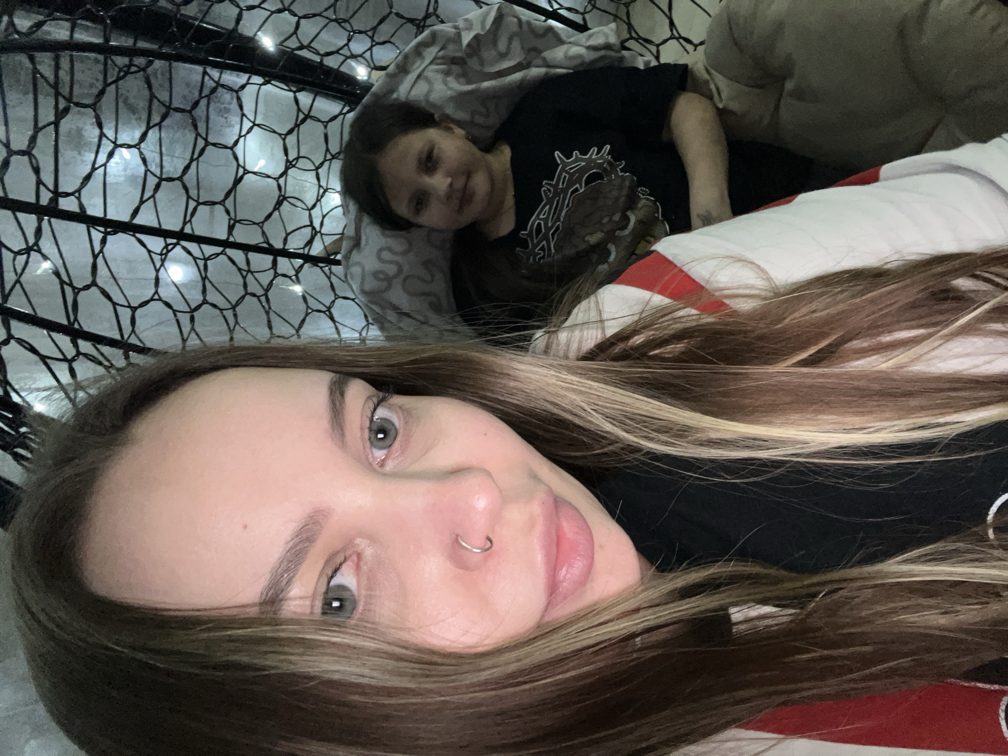 Two individuals are visible. One is a child with dark hair, wearing a black shirt with a graphic design, resting against a gray pillow. The second person, who appears to be taking the selfie, is an adult with long, highlighted hair, wearing a nose ring. They are lying on a white and red patterned surface. The background includes a net-like structure with lights interspersed. The location in the city of Novopokrovskaya, Russia, is not discernible from the image.