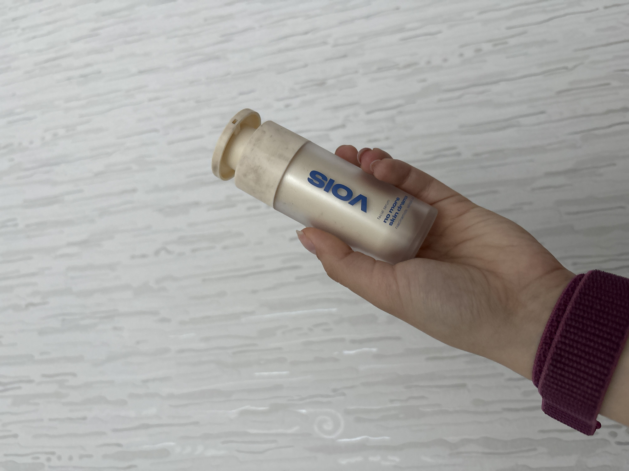 A hand holds a bottle of "Sioa" facial serum against a light-colored wall with a slightly textured, linear pattern. The bottle is a cream color with blue lettering. The hand holding the bottle has fair skin and wears a maroon-colored fabric watch band. The city of Perm, Russia is not evident in the image.