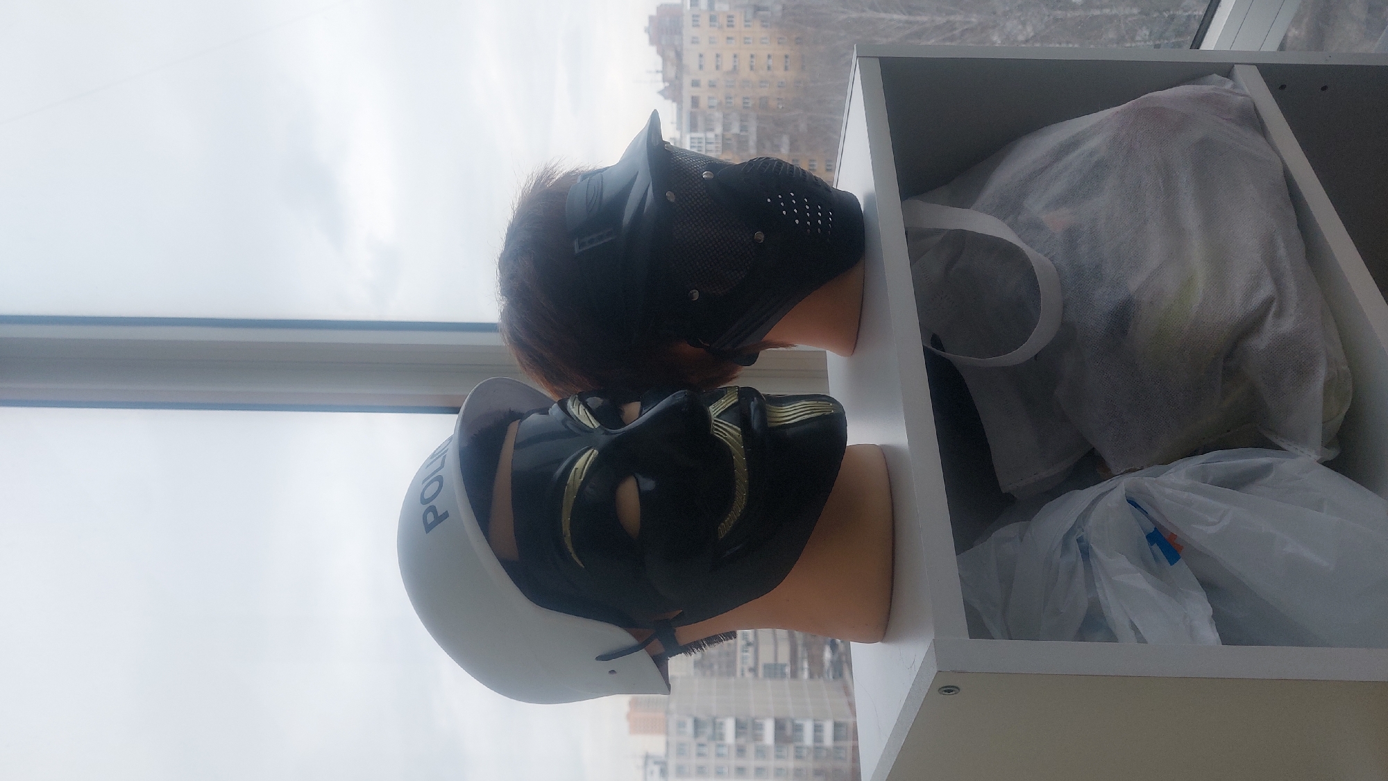 The image captures an indoor scene next to a window, with a view of a multi-story building, indicative of an urban environment, likely in Novosibirsk, Russia. Situated on a white shelf are two mannequin heads. The lower mannequin is wearing a police helmet over a black mask with gold accents. The upper mannequin head has brown hair and is wearing a black, full-face mask with ventilation holes. The shelf also contains a collection of items wrapped in white bags. The overall lighting is subdued, possibly due to an overcast day.