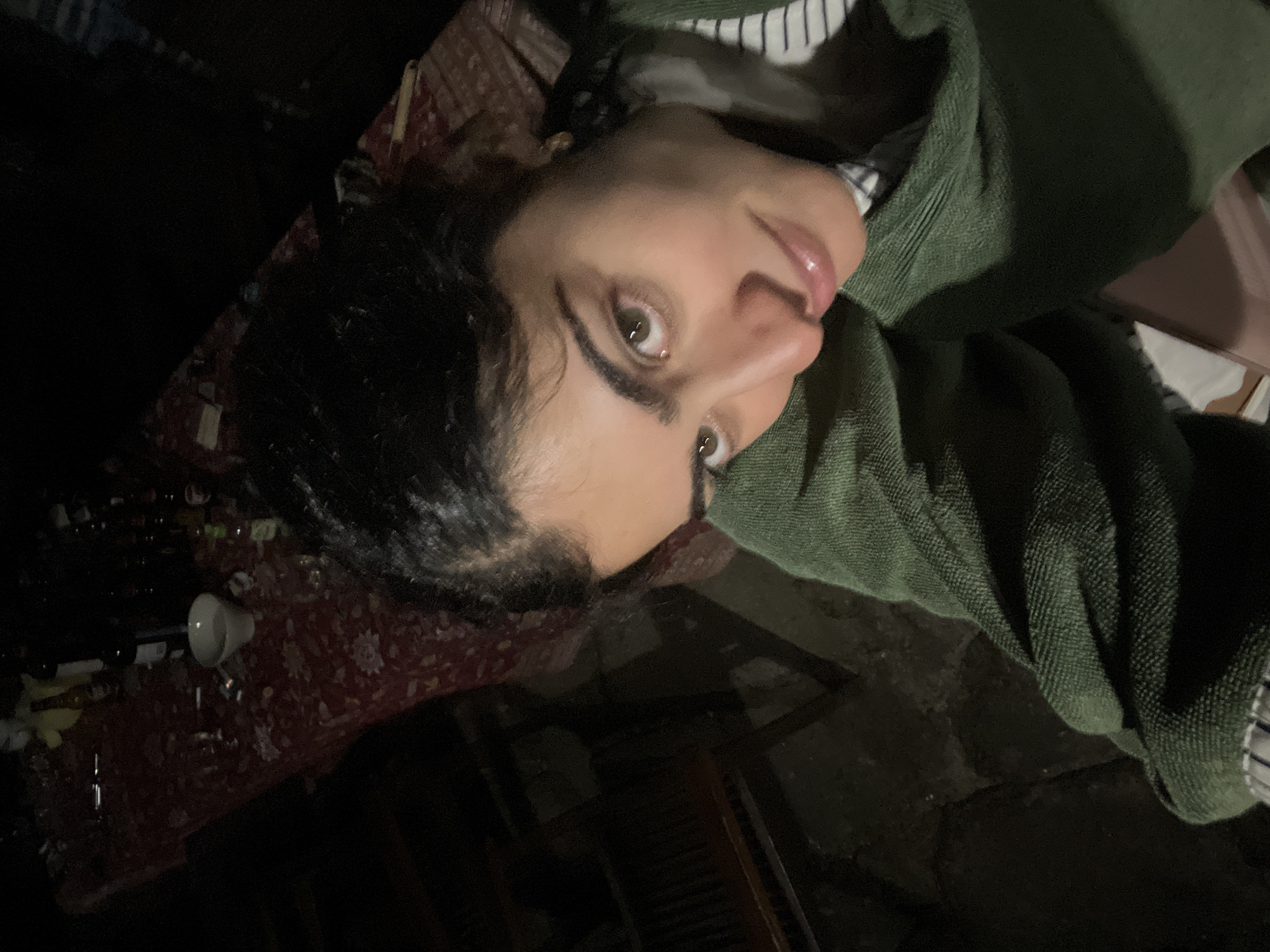 A close-up, high-angle selfie is taken by a young woman with dark hair and eyes. She is wearing a green, textured jacket over a striped shirt. Her head is tilted, and she is looking directly at the camera with a subtle smile. The background is dimly lit, showing a table covered with a red tablecloth. On the table, there are several bottles, a white bowl, and possibly some discarded cigarette butts. The overall environment appears to be outdoors or in a poorly lit indoor space. The location is identified as Felgueiras, Portugal.