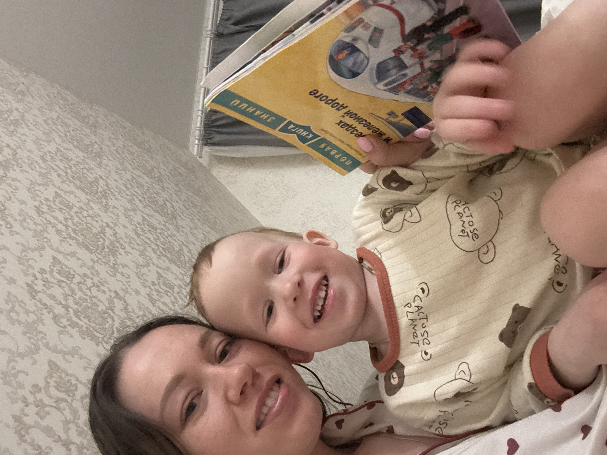 A woman and a child are lying down, posing for a selfie. The woman has brown hair and is smiling. The child, dressed in a bear-patterned pajama, is also smiling and holding a book. The book's cover features an astronaut and the title is written in Russian. The location is a room with patterned wallpaper. Although the book is written in Russian, there is no information on this image which could indicate that the location is the city of Ferma, Russia.