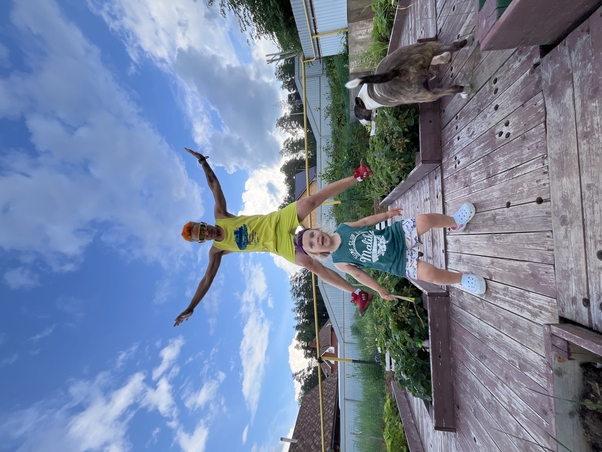 The image depicts an outdoor scene in the city of Chaikovsky, Russia, featuring two individuals and a dog on a wooden deck. In the foreground, a young child wearing a green t-shirt with the text "SURF CLUB Malibu" and patterned shorts stands with a stick in their hand and light blue Crocs. Behind the child, an adult with bright orange hair and yellow sunglasses is captured mid-air, appearing to be jumping. The adult is wearing a yellow tank top and shorts, with red shoes. To the right, a bull terrier dog is facing away from the camera, looking towards a garden area. The background consists of a garden with plants, houses with tiled roofs, a yellow fence, and a blue sky with white clouds. The wooden deck is weathered and shows signs of age.