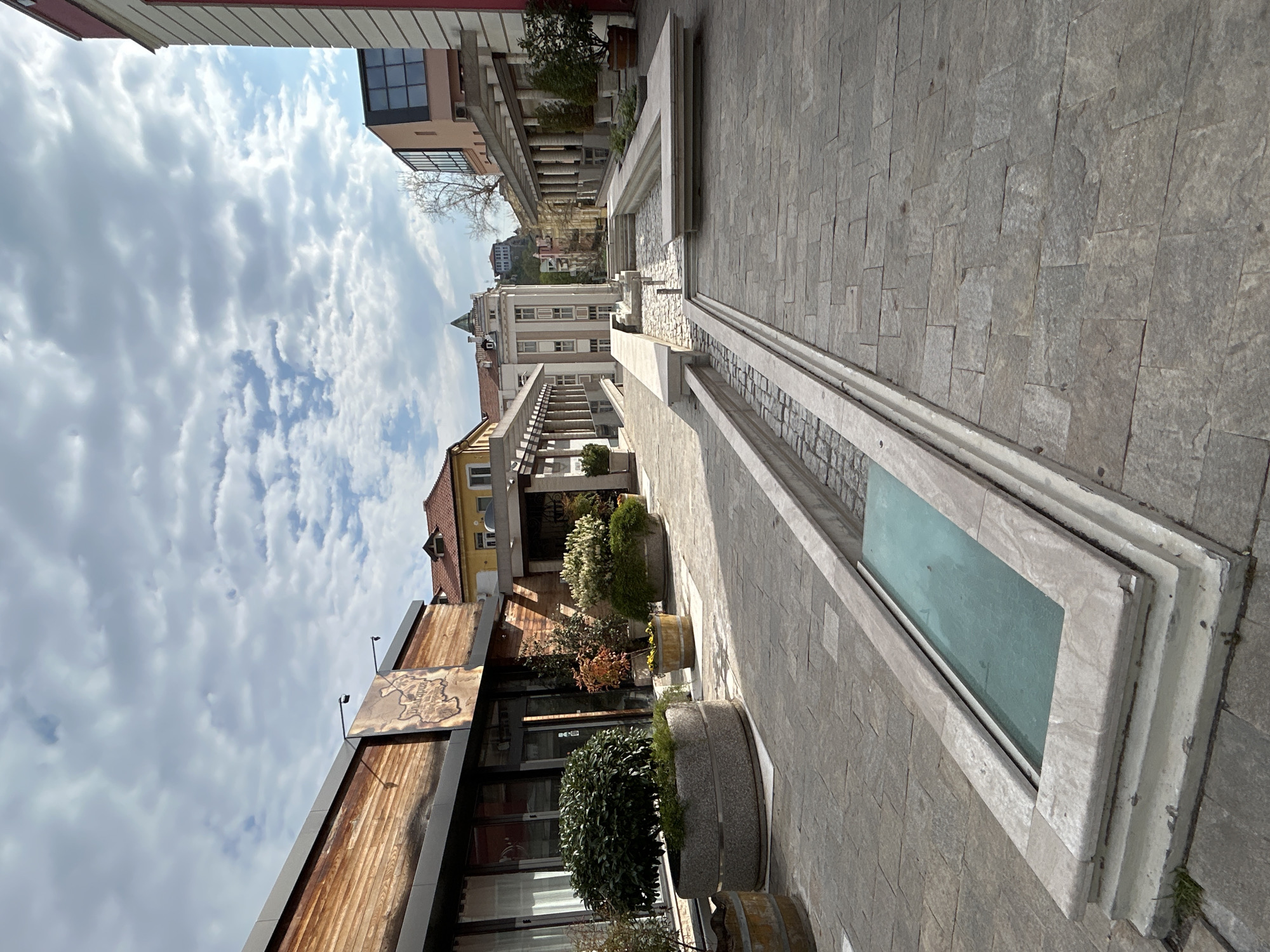 The image shows a street scene in Plovdiv, Bulgaria, featuring a stone-paved walkway flanked by buildings of varied architectural styles. A long, narrow, rectangular structure resembling a water feature or planter runs along the center of the walkway, with a glass-covered section visible. The buildings on either side display different facades, some with wooden paneling and signage, others with visible windows and balconies. Potted plants are placed along the walkway, adding greenery to the urban environment. In the background, other buildings and a cloudy sky are visible. The overall impression is a modern public space within a historic city setting.