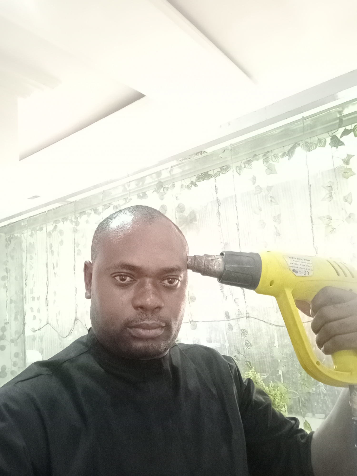 A man, appearing to be in his 30s or 40s with dark skin and a bald head, looks directly at the camera with a serious, unsmiling expression. He is wearing a black collared shirt with a high collar. His right hand holds a yellow heat gun, the nozzle of which is pressed against his left temple, creating a stark and somewhat unsettling pose. Visible text on the heat gun includes "HOT AIR GUN", "POWER", "VOLT", "Hz", and a "CE" mark, though some letters appear mirrored.
The setting is indoors, likely in Gwarinpa, Nigeria, against a large window or glass partition. A light-colored, semi-transparent curtain or screen with a subtle pattern of vines or leaves hangs behind him, partially obscuring the view outside. Beyond the curtain, hints of green foliage suggest an outdoor environment. The ceiling above is white with modern architectural indentations or lighting features, indicating a contemporary interior. The lighting is bright and even, consistent with daytime. The overall impression is a staged photograph, perhaps with an ironic or metaphorical undertone, given the man's direct gaze and the object's symbolic placement.