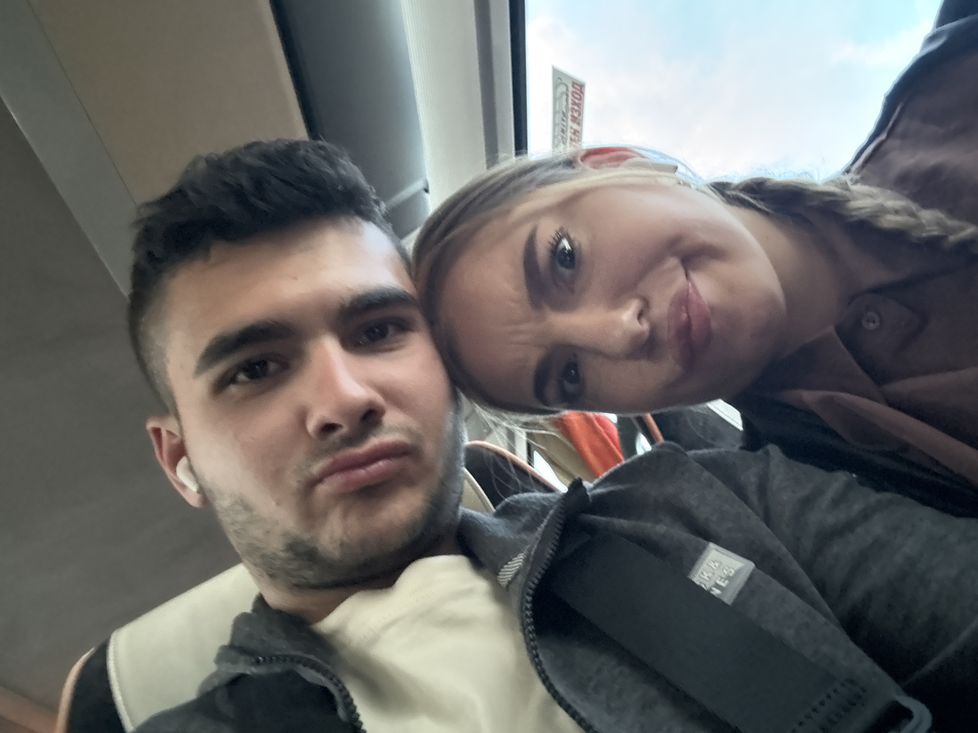 A selfie shows a man and a woman. The man is on the left with short dark hair and a white wireless earbud in his left ear. He is wearing a white shirt and a gray zip-up jacket. The woman is on the right, with blonde hair styled in a braid and wearing a brown shirt or jacket. Their expressions suggest a playful or humorous mood. They appear to be inside a vehicle, possibly a bus or train, given the visible window frame and overhead structure. The exact location within Bouze-lès-Beaune, France, is not discernible from the image alone.