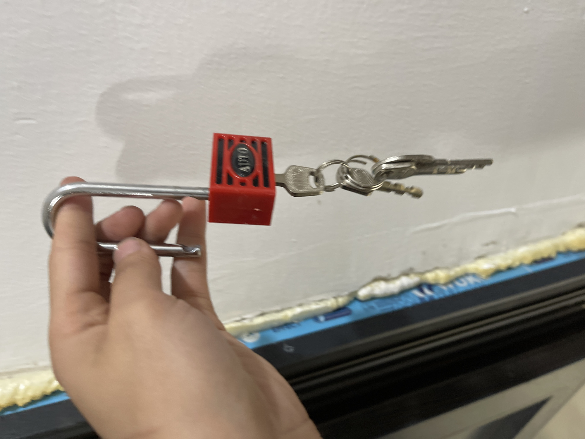 A hand is holding a red padlock with the word "AUTO" visible on it. A set of keys is attached to the padlock. The padlock is hanging against a white wall. To the left of the wall, there is a dark structure, possibly a door or window frame, with some blue and yellow material adhered to it. The scene appears to be indoors. The specific location in Bishkek City, Kyrgyzstan, is not discernible from the provided image.