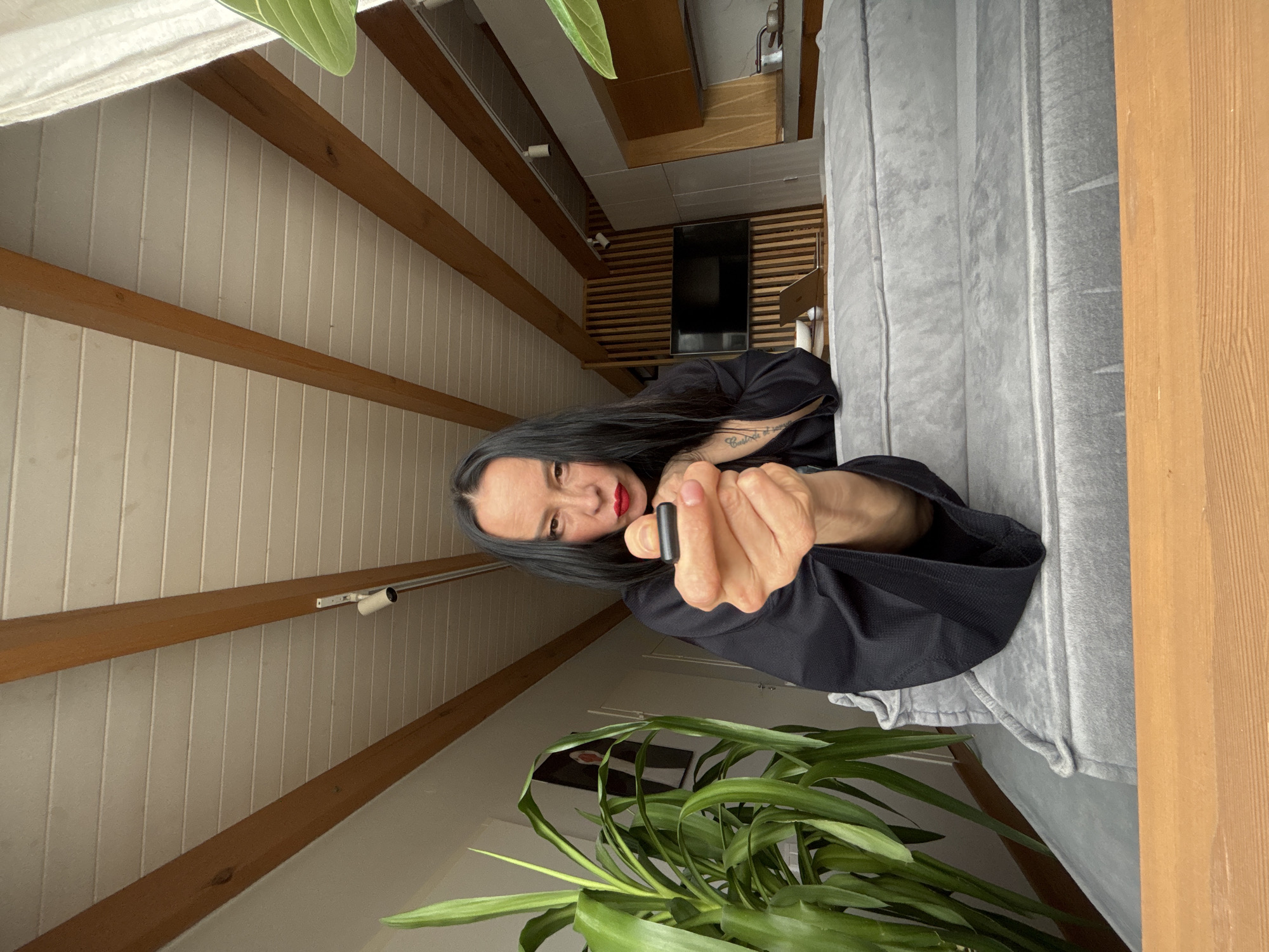 The image depicts a woman in an indoor setting, likely a living room or lounge area, in Chaikovsky, Russia. The shot is taken from a low angle, looking upwards towards the woman and the room's interior.
The main subject is a woman with long, dark hair, wearing a black long-sleeved top or jacket, and bright red lipstick. She looks directly at the camera with a direct and neutral expression. Her right arm is extended towards the viewer, holding a small, black, cylindrical object, which could be a vape pen, remote, or similar device. A tattoo with cursive script is visible on her right forearm, reading "Custodia et..." with the rest of the text obscured.
The room's decor features a modern yet warm aesthetic. On the right, a large grey upholstered sofa or bed is visible. The walls and ceiling are paneled with light-colored vertical wooden planks, intersected by several prominent horizontal wooden beams. In the background, a flat-screen television is mounted on a wall covered with horizontal wooden slats. A small laptop or tablet is also visible near the TV. Track lighting fixtures are mounted on one of the wooden beams. A large potted plant with vibrant green leaves is prominent in the lower left foreground, adding a natural element to the space. The setting suggests a comfortable and stylish home environment. No other discernible text is visible besides the tattoo.