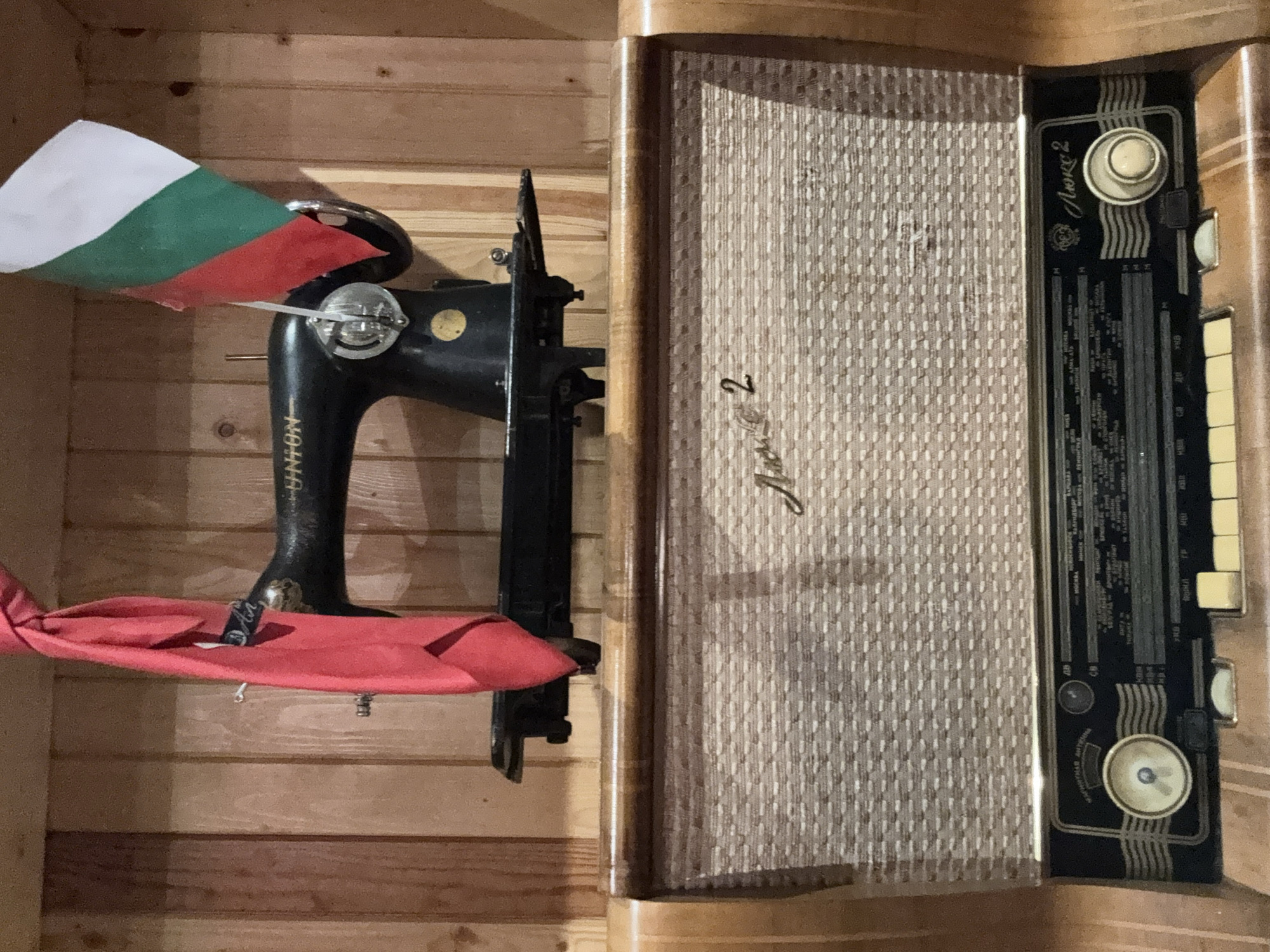 The image shows an indoor scene with two primary objects: an antique sewing machine and a vintage radio. The sewing machine is black and appears to be an older model, potentially hand-operated. It has a Bulgarian flag draped over part of it. A red cloth is beneath it. Next to the sewing machine is a large, wooden-cased radio. The radio has a fabric speaker cover and a control panel with knobs and buttons. Both items are placed inside a wooden structure.
Given the presence of the Bulgarian flag and the "Алко 2" (Alko 2) name on the radio, it's plausible the location is in Bulgaria. Without other information, a location within Sofia, Bulgaria, cannot be conclusively determined from the image.