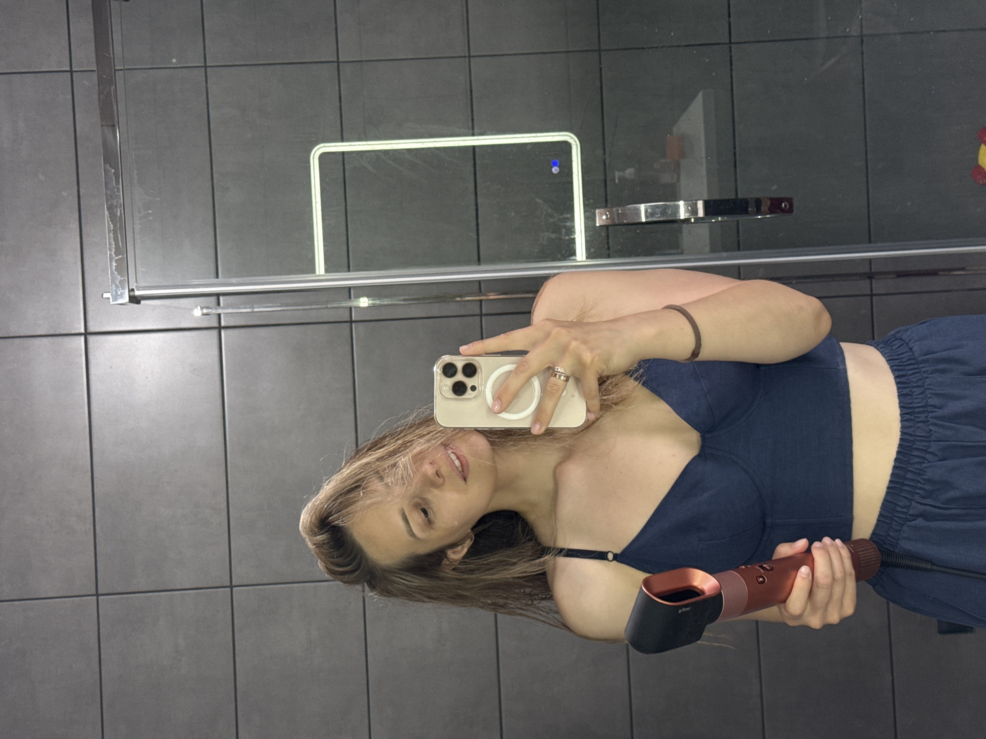 The image is a selfie taken in a bathroom, likely inside a building in the city of Chaikovsky, Russia. The room has grey, square-tiled walls. A woman is holding a cell phone and a Dyson hair dryer, apparently drying her hair. She is wearing a blue bra and dark blue sweatpants. The reflection in the bathroom mirror shows a chrome shower door frame.