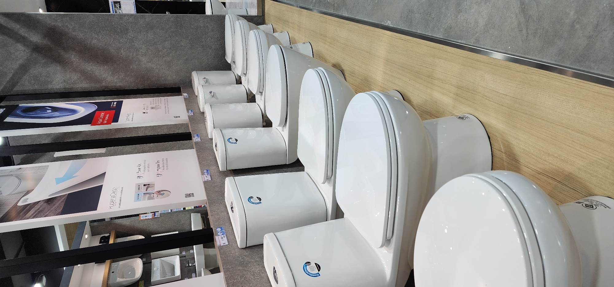 The image shows a retail display of white ceramic toilets in a store, possibly a home improvement or bathroom fixture store. Several toilets are lined up on a wooden platform, with a textured gray wall in the background. Some toilets have their tanks attached, while others appear to be just the bowls. There are also some promotional posters or display boards behind the toilets with graphics and text, some of which mention "Hygiene Clean" and "Aqua Ceramic". The lighting appears to be artificial and the scene is well-lit, with no visible people. The overall impression is a clean and organized product display. The location is specified as Baan Chaweng, Thailand.