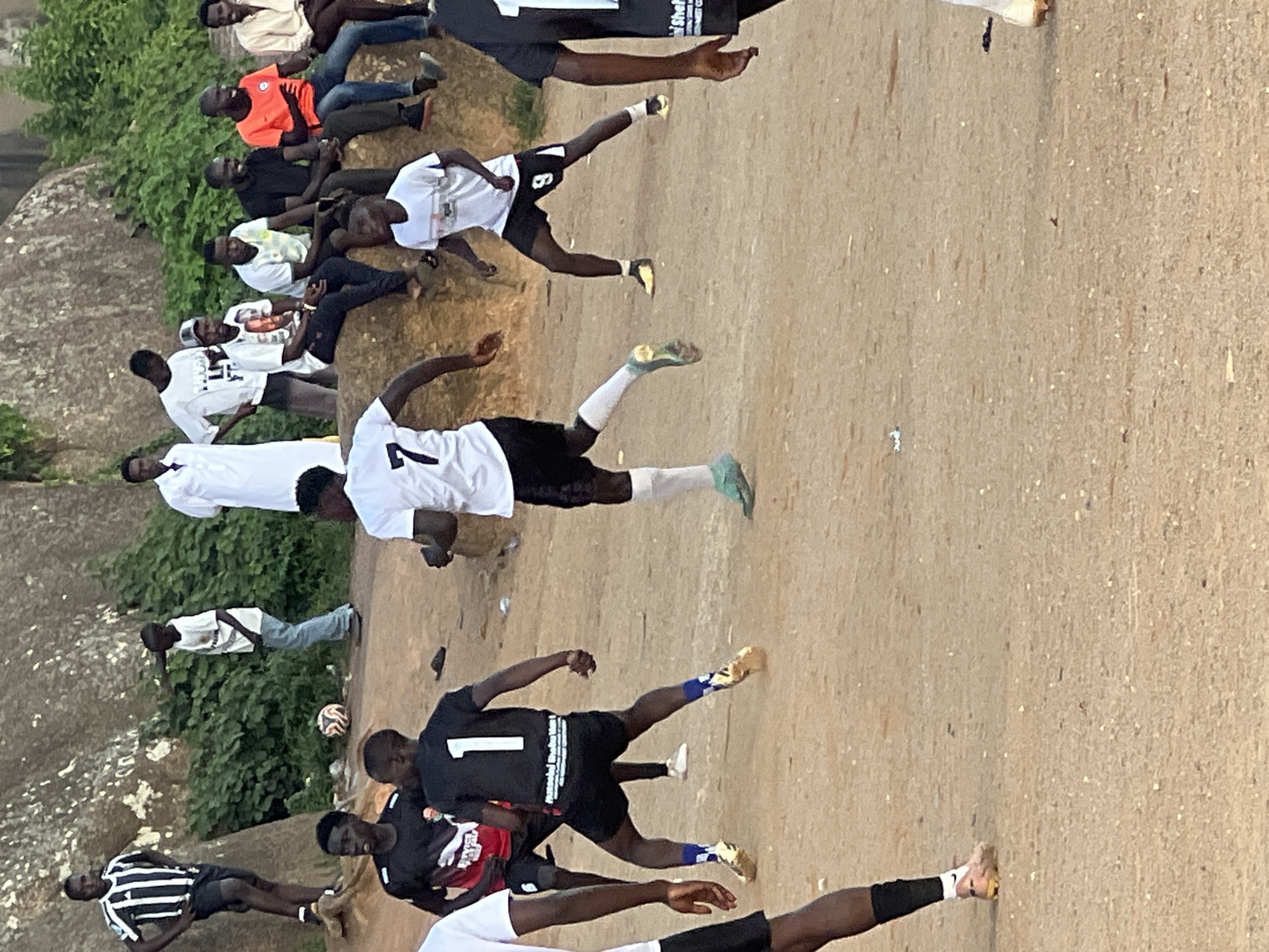 The image depicts a group of young men playing football on a dusty, uneven field, likely in the city of Jos, Nigeria, given the rocky, arid landscape and the attire of the individuals. In the foreground, two players in black jerseys with white numbers "1" and "7" are actively engaged in the game. The player with number "1" is wearing black shorts and blue socks, while the player with number "7" is wearing black shorts and white socks. Another player in a black and red jersey is partially visible in the left foreground. In the background, several men are seated on a rocky ledge, observing the game. One man in a white robe stands near a large rock formation covered in green vegetation. The overall scene suggests a casual, local football match taking place in an outdoor environment.