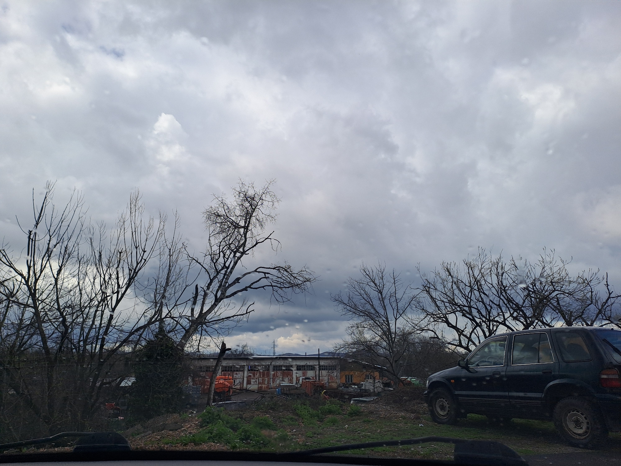 The image shows a drab, overcast scene, possibly taken in or near the city of Varshets, Bulgaria, given the user's instruction. In the foreground, a dark-colored SUV is parked on uneven ground, its side facing the viewer. The terrain is a mix of dirt, sparse green vegetation, and bare patches. Several leafless trees are scattered throughout the scene, their bare branches creating a network against the gray sky. Behind the trees, a dilapidated, single-story industrial building is visible. The building appears to be constructed of corrugated metal and concrete, with numerous window openings. Orange safety vests are piled in front of the building along with other debris and construction materials. The sky is uniformly gray and overcast, filling the majority of the image. The photo seems to be taken from inside a vehicle, with a portion of the dashboard visible at the bottom of the frame. There are no people visible.