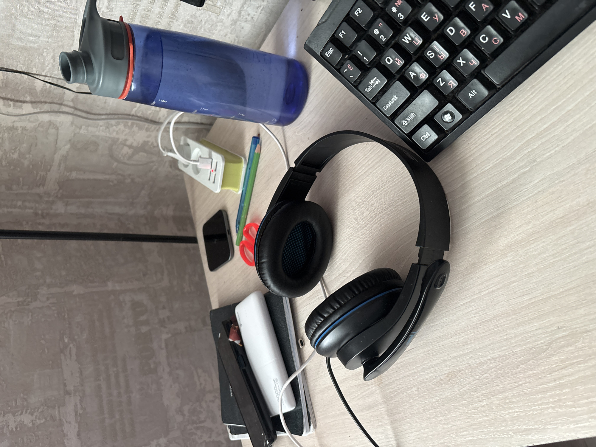 This is a close-up, eye-level shot of a desk with various items on it. The lighting is natural, coming from the left, and casts soft shadows. The background is a textured wall, possibly wallpaper, with vertical lines. The desk surface is a light-colored wood grain laminate.
Dominating the foreground is a pair of black over-ear headphones with blue accents. They are positioned as if just placed down, with the headband extended and the earcups resting on the desk. To the right of the headphones is a black computer keyboard, with its keys visible, including "Esc", "F1", "F2", "Tab", "Caps Lock", "Ctrl", "Alt", and letters from the Russian alphabet, indicating a Cyrillic layout.
Behind the headphones, to the left, is a stack of objects. The top item appears to be a black smartphone lying on its side. Beneath it is a white power bank with some text visible, and below that, a black notebook or binder.
Further back on the desk, towards the center, is a yellow power strip with two white electrical outlets. A white USB cable is plugged into one of the outlets, and a red light is visible on the power strip, suggesting it is active. To the left of the power strip, a black object, possibly another phone or a small electronic device, is placed on the desk.
To the right of the power strip and slightly behind it is a blue translucent water bottle with a grey lid and an orange ring around the neck. The bottle contains a clear liquid, likely water.
In the middle ground, between the notebooks and the water bottle, are a red pair of scissors and a green pencil.
The scene suggests a workspace or a bedside table, possibly in a dorm room or an office. There are no people visible in the image.
The image does not provide any information or visual cues to determine its location in the city of Sochi, Russia.