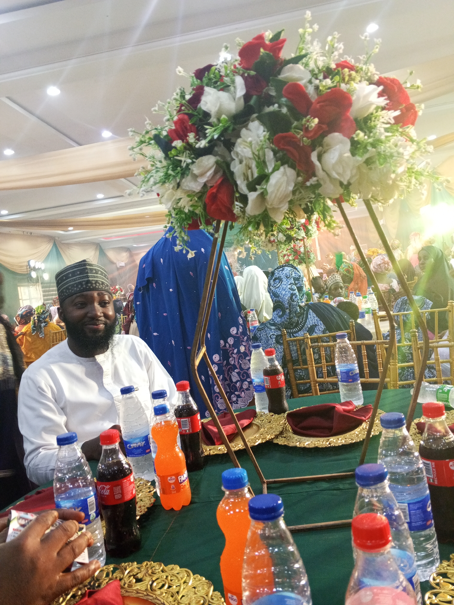 The image shows a gathering of people indoors, likely at a celebration or event. A man in the foreground, wearing a white traditional garment and a patterned cap, smiles warmly. He is seated at a green-covered table with several bottles of beverages, including Coca-Cola, Fanta, and bottled water. The table is decorated with ornate golden platters and red napkins.
Behind the man, a large floral arrangement with red and white flowers stands prominently, supported by golden poles. Further in the background, other guests are visible, some wearing colorful attire and headscarves. A woman in a striking blue garment with intricate patterns is also seated at a table. The setting appears to be a banquet hall, with draped fabric and lighting fixtures visible on the ceiling, suggesting an evening or indoor event. The overall mood seems festive and convivial.
Visible text on the beverage bottles includes "Coca-Cola," "FANTA N300," and "CWAY."