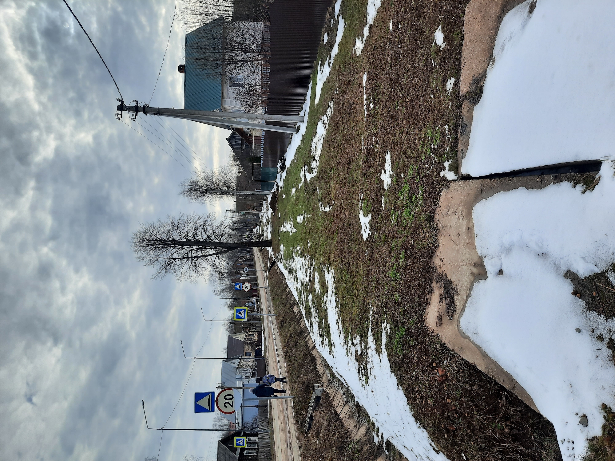 This image captures a street scene in what appears to be a semi-urban or rural settlement during late winter or early spring, consistent with the given location context of Krasnogorskoye, Russia. The sky is heavily overcast with grey clouds, indicating a cloudy daytime.
In the foreground to the right, patches of melting snow are visible on rough, brown ground interspersed with sparse green grass, next to a concrete or stone ditch that also contains snow. The middle ground features a road or wide track, running roughly parallel to the viewer, where two figures, an adult and a child, are walking away. They are positioned on a sidewalk or shoulder to the left of the road.
Several pieces of street infrastructure are prominent. Multiple utility poles with power lines crisscross the frame, alongside streetlights. Road signs are clearly visible: a circular "20" sign, indicating a speed limit, and a blue triangular sign with a pedestrian icon, marking a pedestrian crossing.
In the background, beyond the road and sidewalk, various houses and buildings, some partially obscured by fences, line the street. Many trees are bare, reinforcing the late winter or early spring setting. The overall scene suggests a quiet, everyday moment in a residential area.