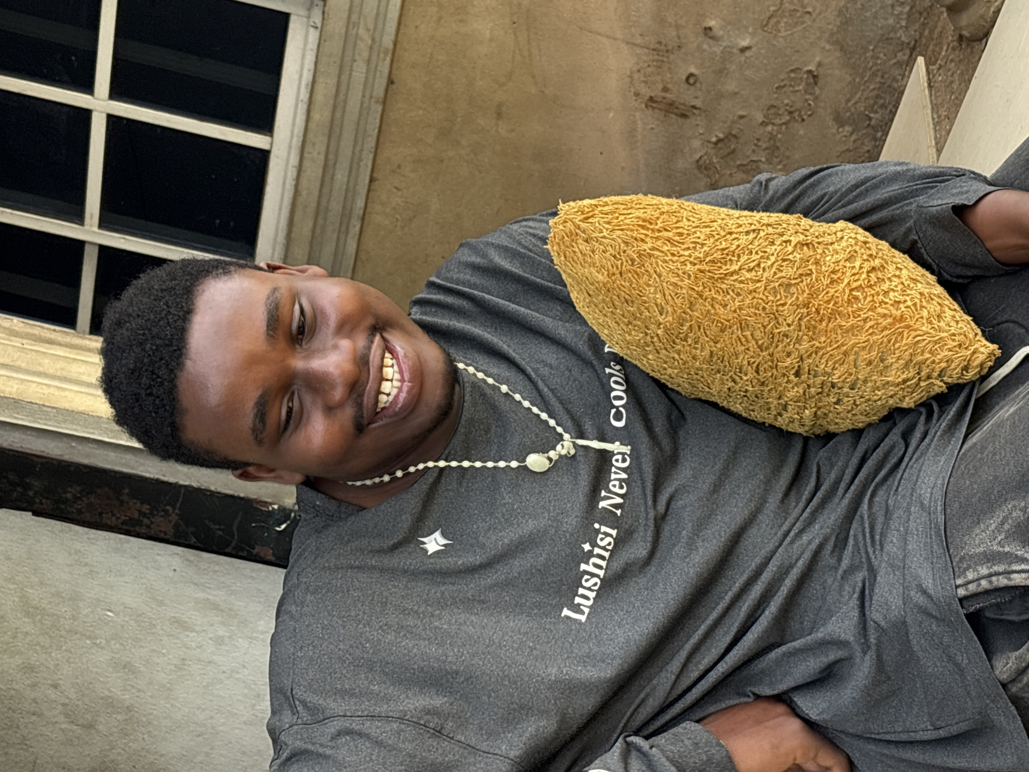 A young, dark-skinned man is smiling at the camera. He is wearing a gray t-shirt with white text ("Lushisi Never Cools") and a white beaded necklace. He is holding a yellow, textured object that appears to be a pillow or cushion. The background includes a window with multiple panes and a beige wall. The location is Ikot Ekpene, Nigeria.