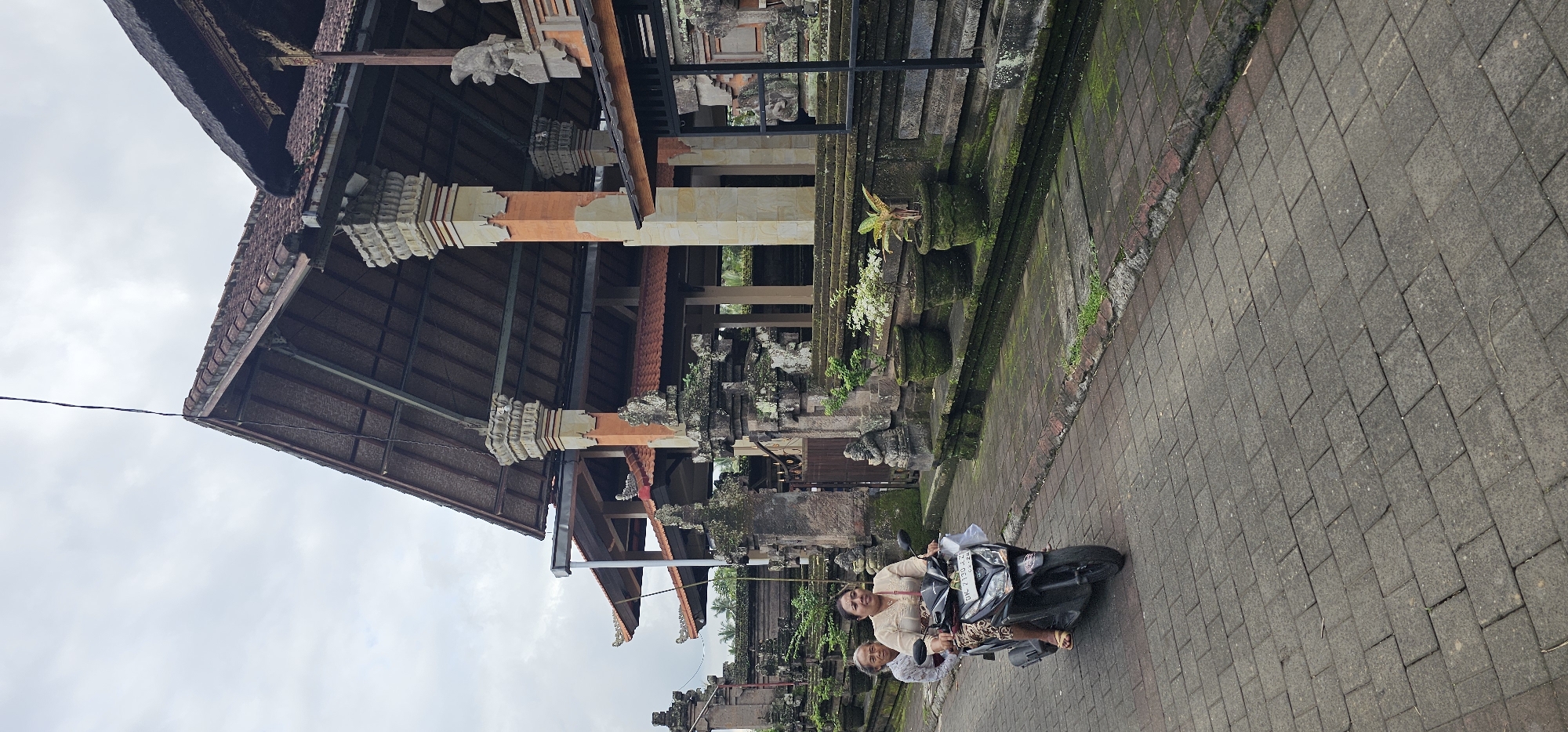 The image shows a traditional Balinese structure in Ubud District, Indonesia. The building features multiple levels, a steeply pitched roof with thatched sections, and ornate stone carvings. It's built with a combination of stone, wood, and brick, exhibiting a weathered appearance with moss and signs of age. The foreground features a paved area, possibly a walkway or street, and a woman rides a scooter with another woman as a passenger, with license plate "DK 2732 AK" visible. The overall environment suggests a historical or cultural site within a tropical climate. The sky is overcast.