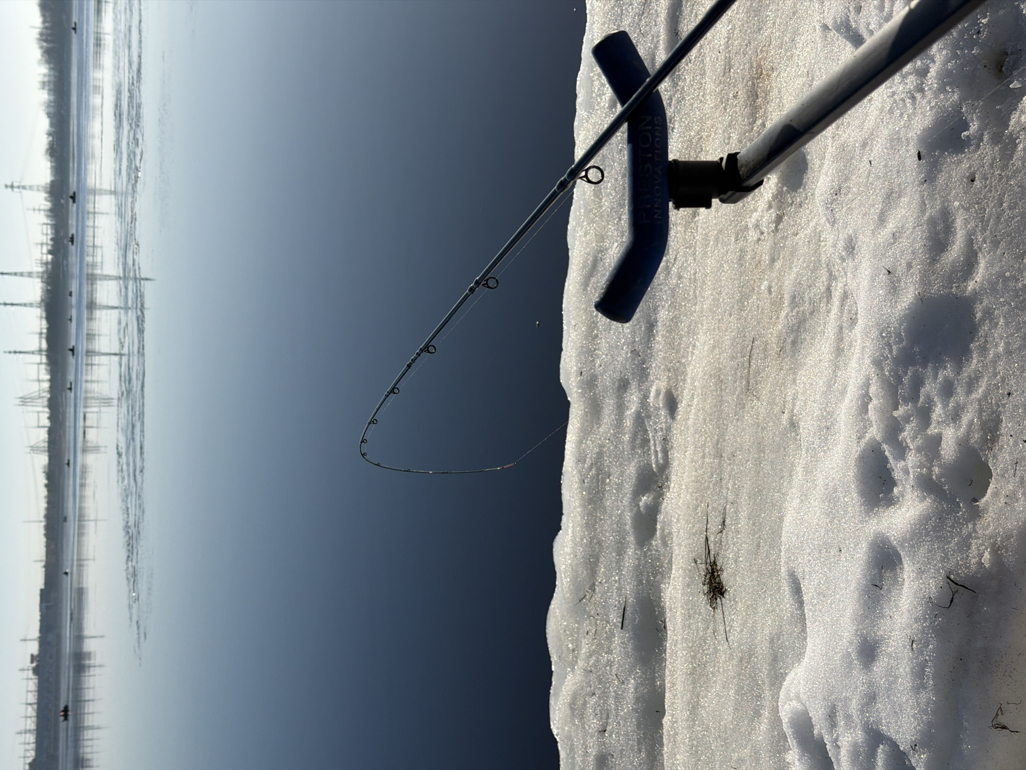 The image shows a fishing rod set up on a snowy bank, with a body of water visible in the background. The rod is supported by a blue rod rest marked "PRESTON INNOVATIONS." The foreground is covered in snow. The water appears dark and reflective. In the distance, there are structures that look like power lines or industrial towers.
Without more context, pinpointing the precise location within Chaikovsky, Russia, is not possible. However, the presence of the industrial structures suggests the location could be near a body of water (likely the Kama River) with industrial infrastructure nearby. The scene suggests a winter fishing activity.