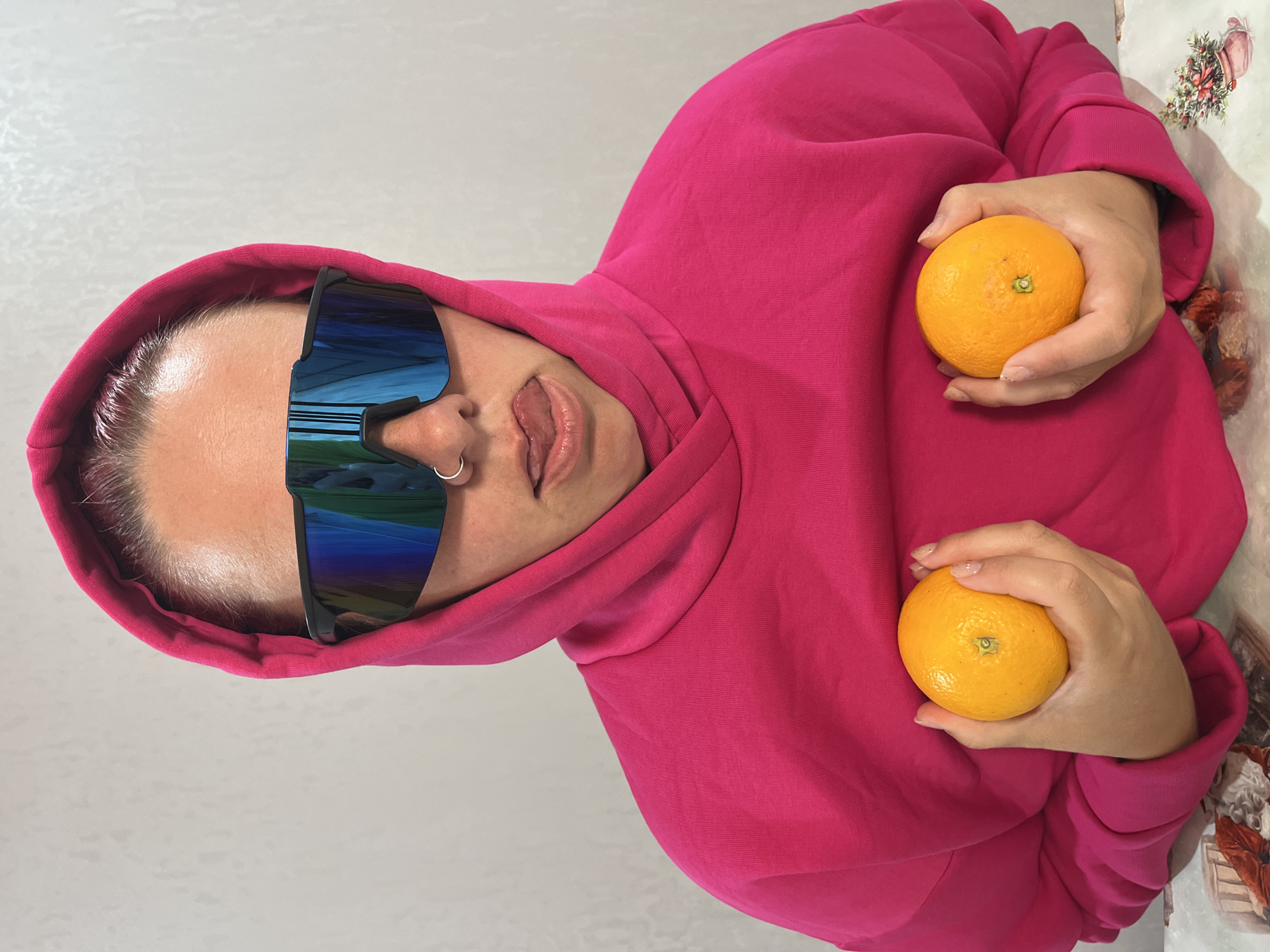 A person wearing a bright pink hooded sweatshirt and large, reflective sunglasses is holding an orange in each hand. They are sticking their tongue out. The setting is indoors, with a neutral-colored wall in the background to the left and a patterned surface to the right. The image appears to be a snapshot or self-portrait. The location, Chaikovsky, Russia, cannot be confirmed from visual data.