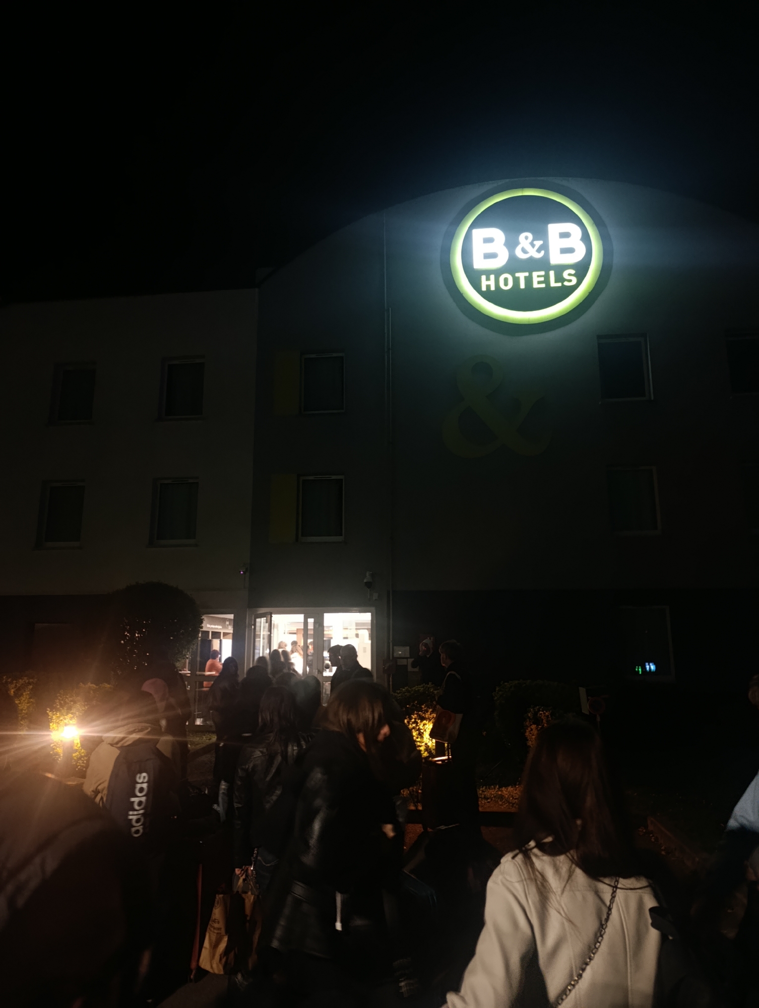 The image shows the front entrance of a B&B Hotel at night. The hotel's illuminated sign is visible. A group of people are gathered near the entrance, some appearing to be entering or exiting. The location is likely Betting, France. Details about specific individuals or interactions within the group are limited due to the lighting and perspective.
