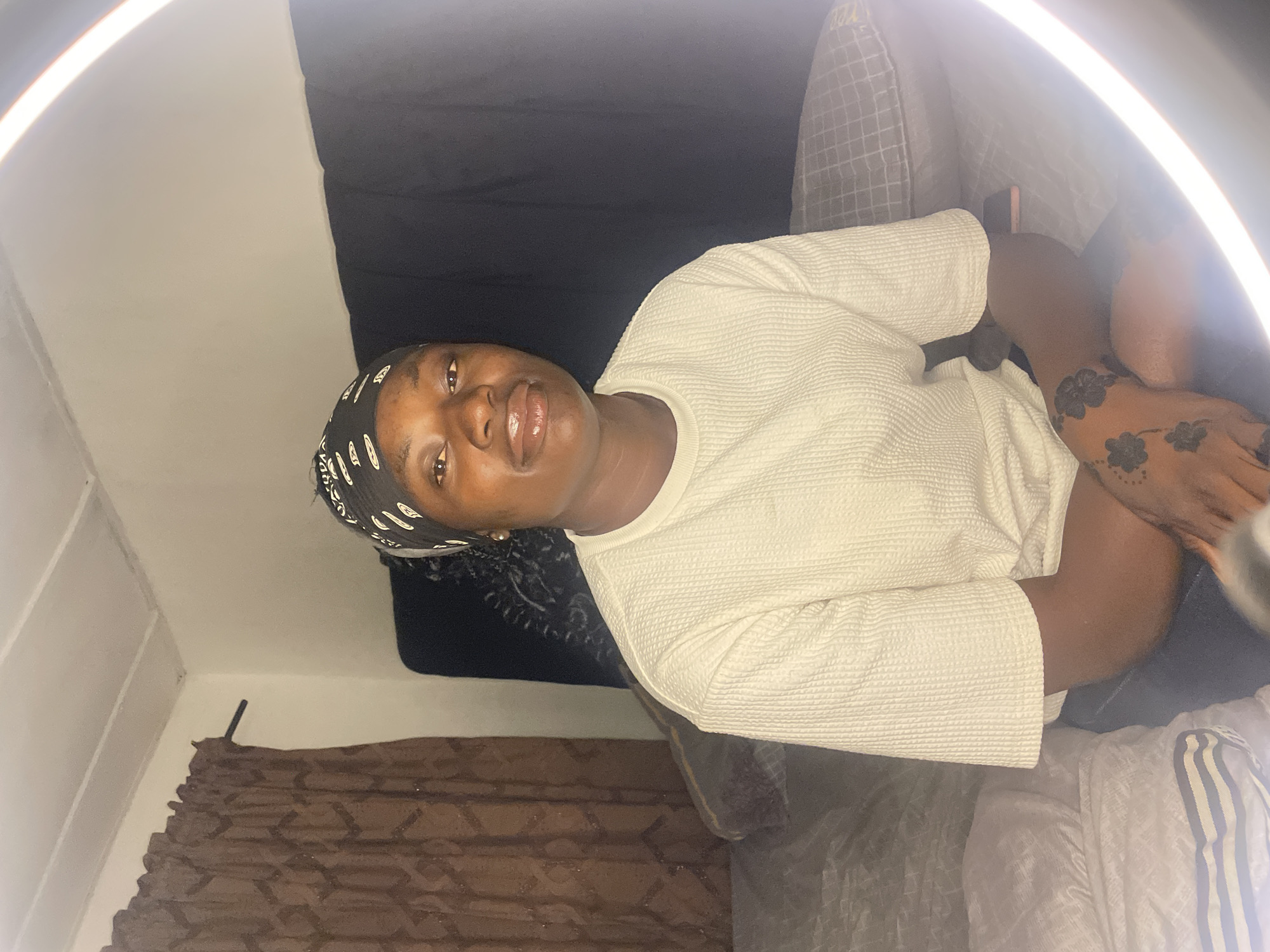 A medium shot depicts a young Black woman indoors. She is wearing a patterned headscarf and a textured, off-white, long-sleeved top. Floral tattoo designs are visible on her hand. A ring light is positioned in the upper right. The background includes a white wall, a black drape, and patterned curtains. While the image provides details about the subject and setting, the location within the city of Jos, Nigeria cannot be confirmed based solely on the visual information.