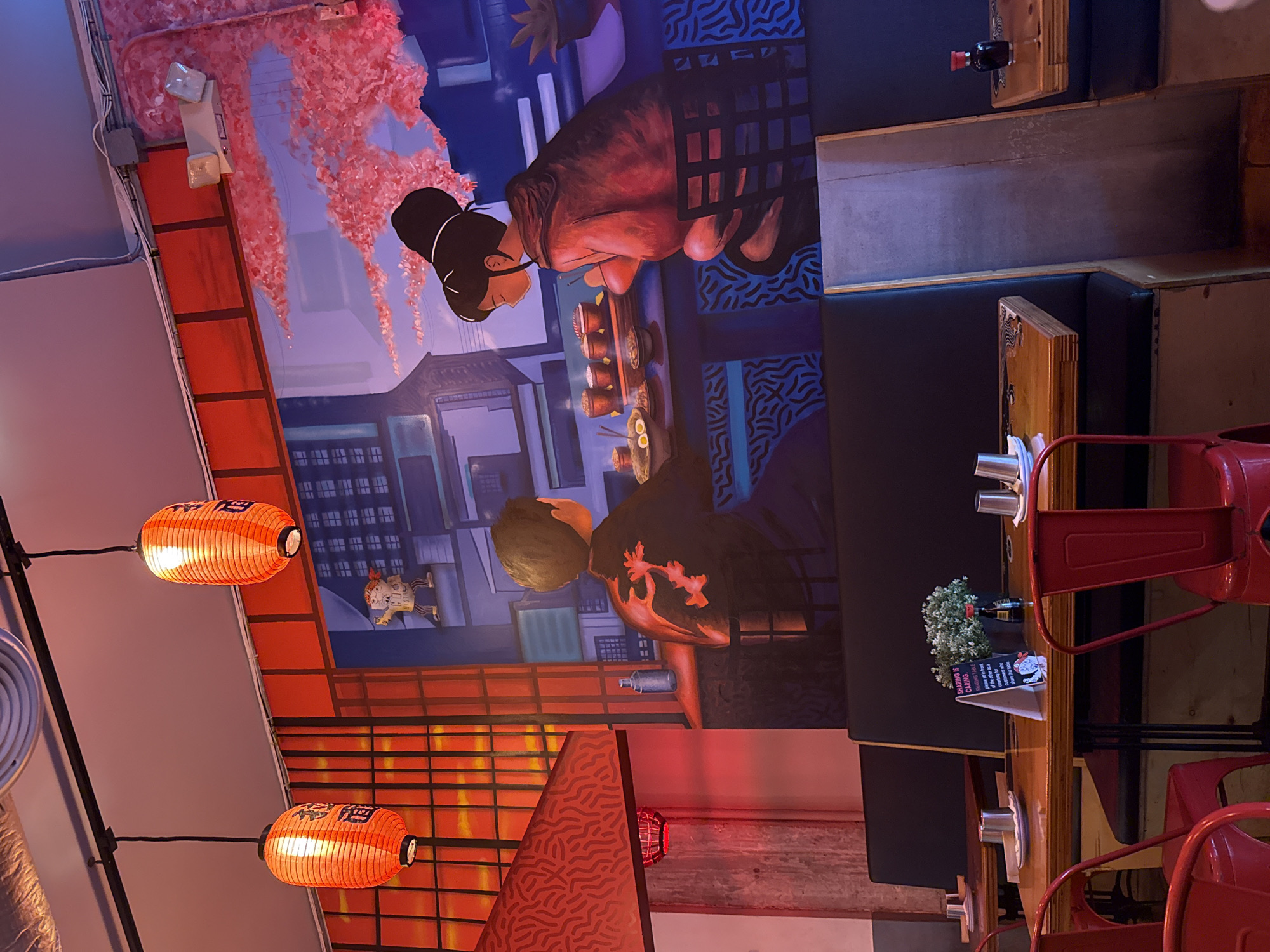 The image captures the interior of a Japanese or Asian-themed restaurant, featuring a vibrant mural, traditional lanterns, and dining furniture.
The central and most prominent feature is a large wall mural depicting a stylized, anime-like city scene. It shows traditional Japanese buildings under a blue sky, with cascading pink cherry blossoms dominating the upper left. Two main figures are part of the mural: on the left, a female figure with a black bun, glasses, and dark attire, appears to be holding chopsticks over a bowl of food. To the right and below her, a male figure with a light haircut is shown from the back, wearing a black garment with an orange/pink pattern. Both are positioned around a table with multiple small dishes, suggesting a meal in progress. A smaller, cartoonish figure is visible on a ledge in the mural's lower-left corner.
Two illuminated orange paper lanterns, adorned with black Japanese characters, hang from the ceiling on black wires in the foreground. The restaurant is furnished with dark upholstered booth seating along the wall and wooden tables. Bright red metal chairs with slatted backs are pulled up to the tables. On one table, a small vase with white flowers, a dark bottle (possibly soy sauce), and a small white triangular sign are visible. The sign reads, "SHARING IS CARING: Sit on one side of the table for conversation & convenience. Sit on the other as it creates a private space, for diners who share this table." Below the text is a small drawing of a figure holding chopsticks.
The lighting is dim, indicating an evening setting, with artificial light source from the lanterns and industrial-style wall lights on the left. The ceiling reveals an air vent and exposed conduit. The overall atmosphere is modern yet infused with traditional Japanese cultural elements.