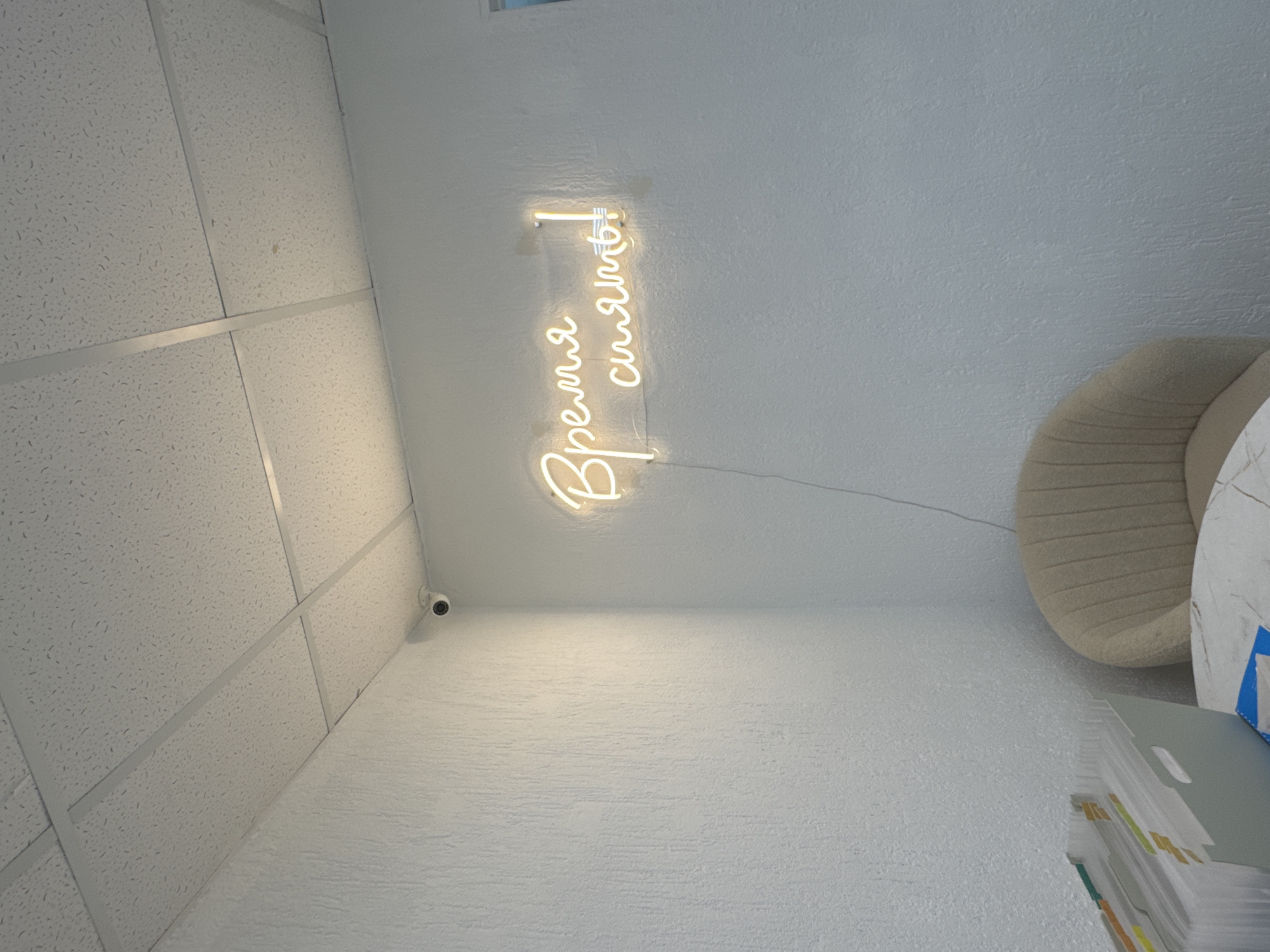 A neon sign displaying "Время сиять!" which translates to "Time to shine!" in Russian, is affixed to a textured white wall in a room. To the upper left, a small, circular security camera is mounted on the ceiling. The ceiling itself is a white, suspended tile system with metal grid supports. In the lower right, a portion of a beige upholstered armchair is visible. In the lower left, a grey organizer filled with folders and documents sits on a table with a marbled surface. The lighting appears to be a mix of overhead fluorescent lights and the glow from the neon sign. The overall impression is of an indoor space, possibly an office or a salon, located in the city of Chaikovsky, Russia.